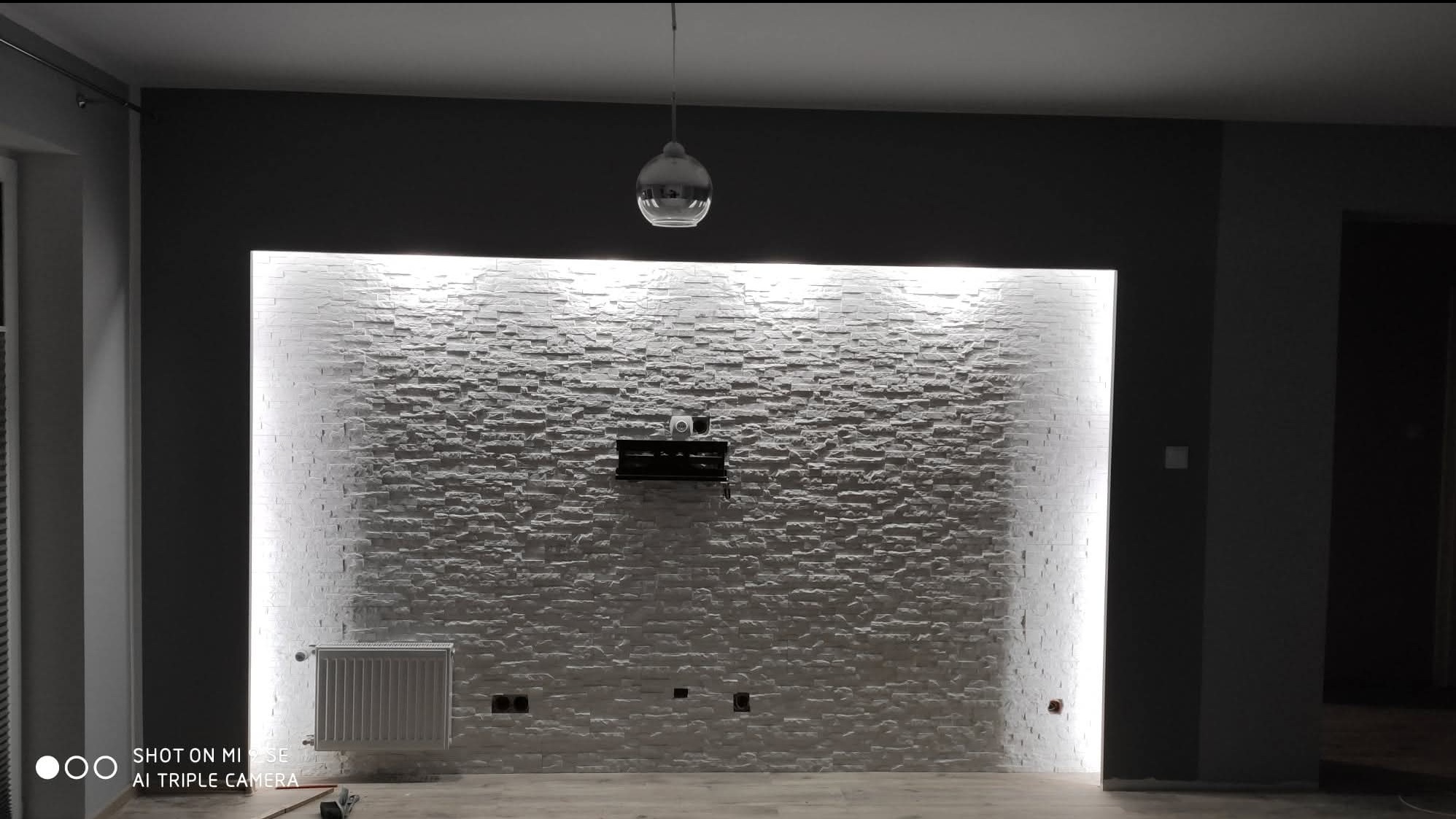 Ściana z dekoracyjnej białej cegły, podświetlona pionowymi pasami LED, z zamontowanym uchwytem TV i wiszącą lampą. Szare ściany boczne i grzejnik dopełniają wystrój.