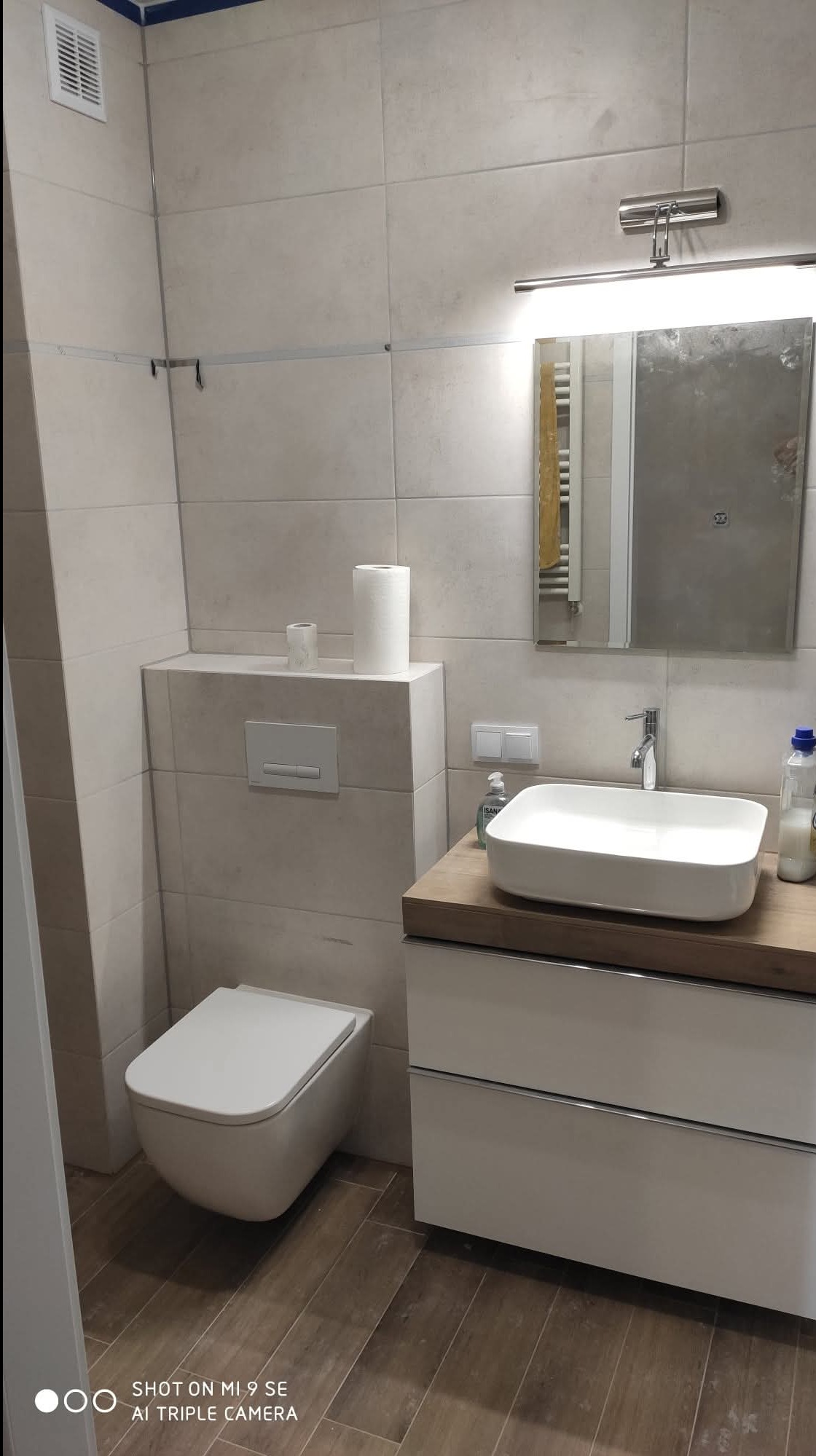 Nowoczesna łazienka z podwieszaną toaletą, umywalką na szafce z drewnianym blatem i lustrem z oświetleniem. Beżowe płytki na ścianach i brązowe na podłodze.