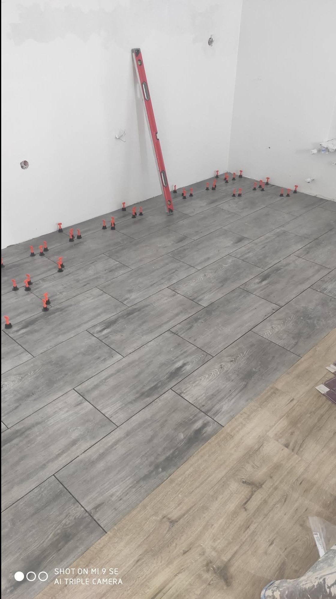 Ułożone szare płytki podłogowe imitujące drewno, z czerwonymi klipsami poziomującymi i poziomicą przy ścianie. Część podłogi jeszcze nie wyłożona.