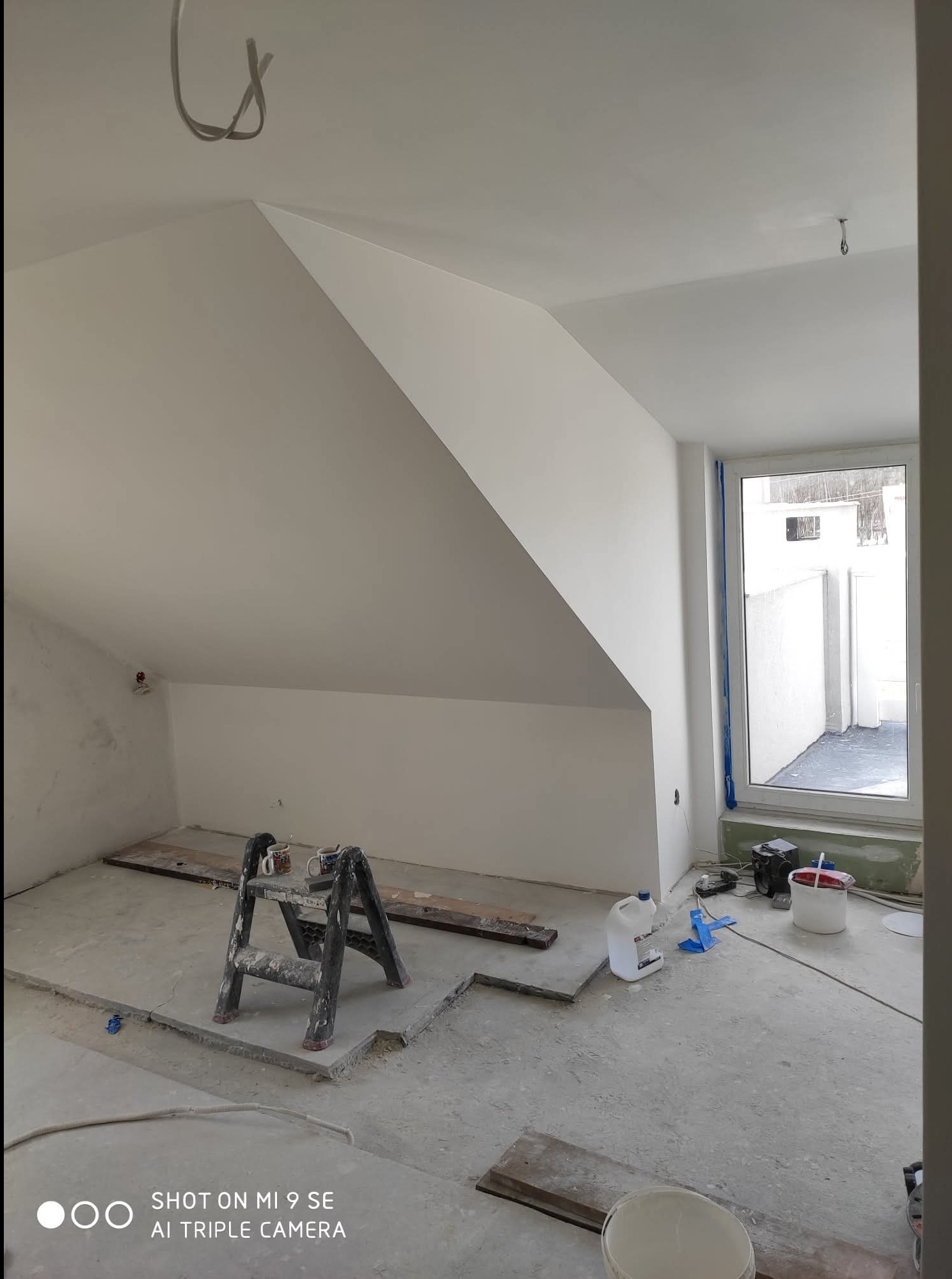 Poddasze po szpachlowaniu i malowaniu na biało, widoczna drabina malarska, okno i przygotowane podłoże. Ściany i sufit pomalowane na jednolity, jasny kolor.
