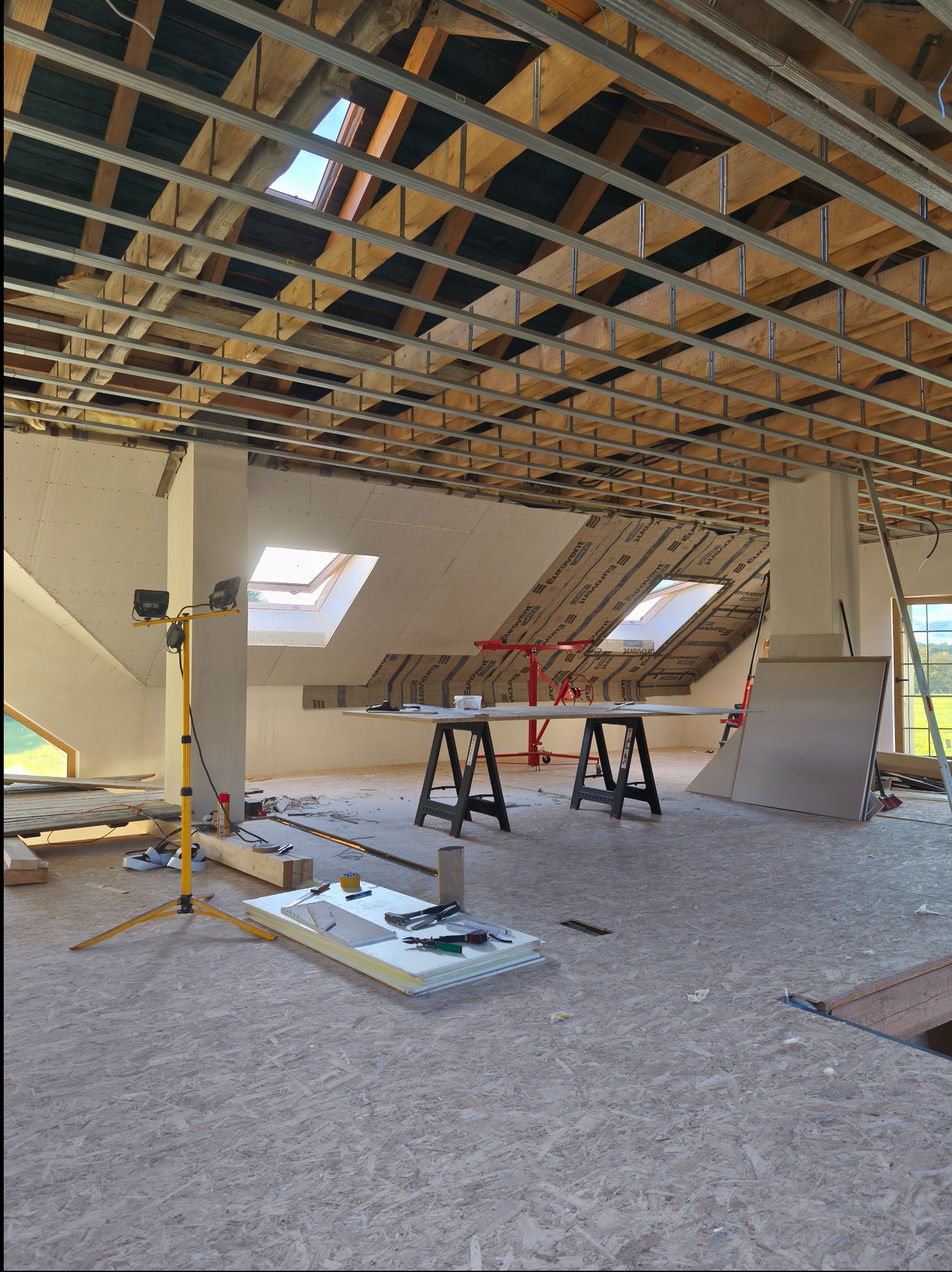 Poddasze w trakcie adaptacji: widoczne krokwie, metalowa konstrukcja sufitu, ocieplenie, okna dachowe, narzędzia i materiały budowlane. Prace wykończeniowe w jasnym wnętrzu.