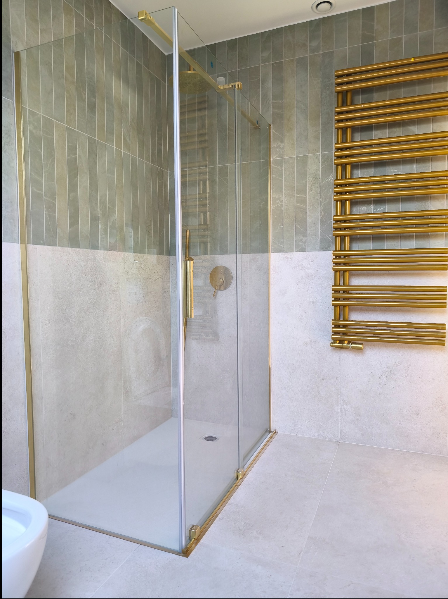 Nowoczesna kabina prysznicowa z złotymi detalami, grzejnik drabinkowy w kolorze złota, płytki w odcieniach szarości. Minimalistyczny design łazienki.