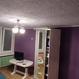 Finish - Fioletowa ściana w pokoju z oknami, grzejnikiem, stołem z telewizorem i szafą z lustrem. Sufit z fakturą. Widok na wnętrze po odświeżeniu.