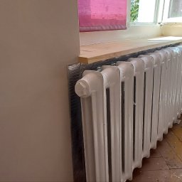Finish - Biały, żeliwny grzejnik po renowacji, zamontowany pod oknem z drewnianym parapetem. Widoczne ślady prac remontowych na podłodze i świeżo pomalowana ściana.