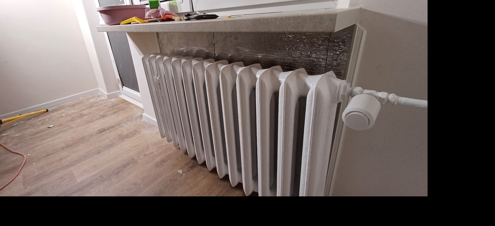 Biały, żeliwny grzejnik po renowacji, z nowym termostatem, na tle ściany z zabezpieczeniem termicznym. Widoczne fragmenty podłogi i okna. Renowacja grzejników.
