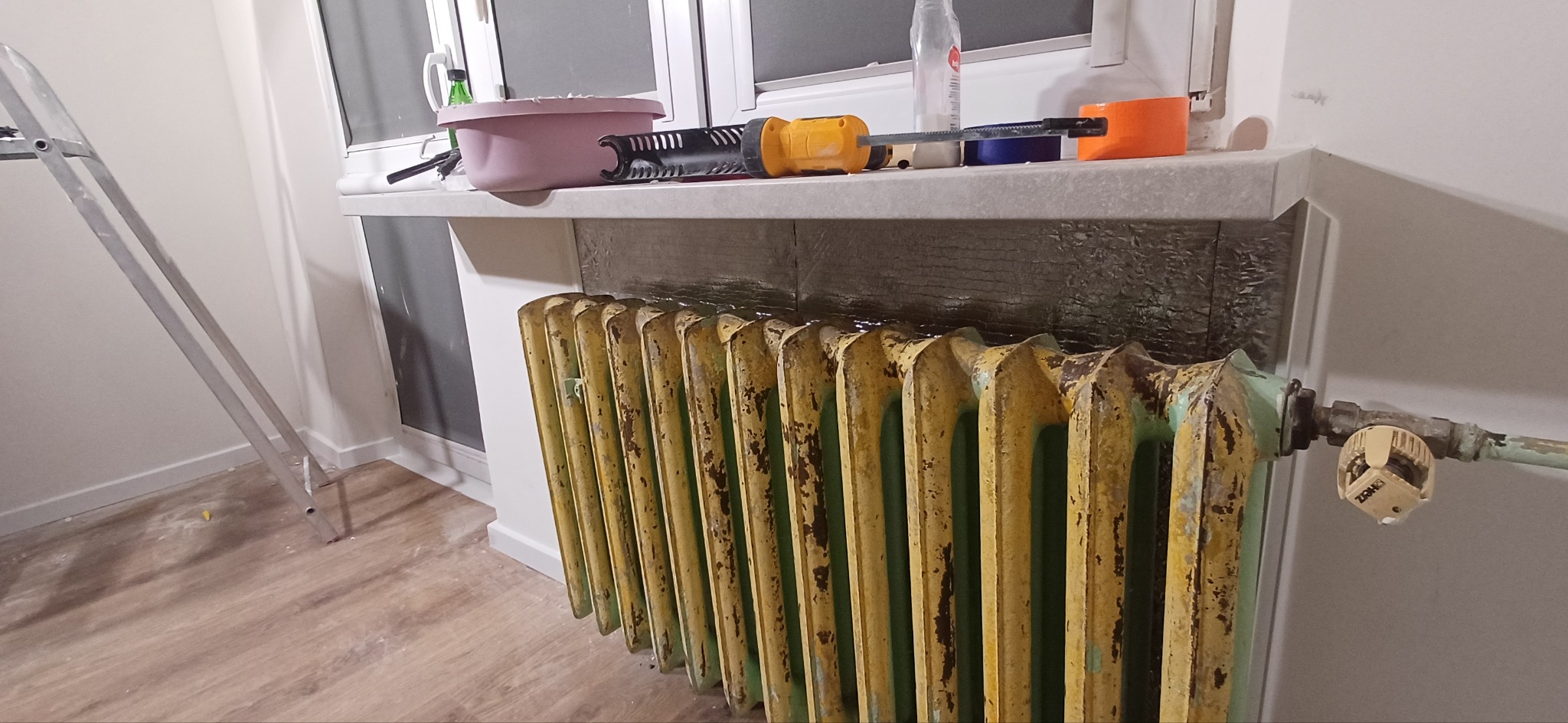 Żeliwny, żeberkowy grzejnik w trakcie renowacji, zabezpieczony folią, z narzędziami na parapecie i drabiną w tle. Widoczne ślady rdzy i starej farby.