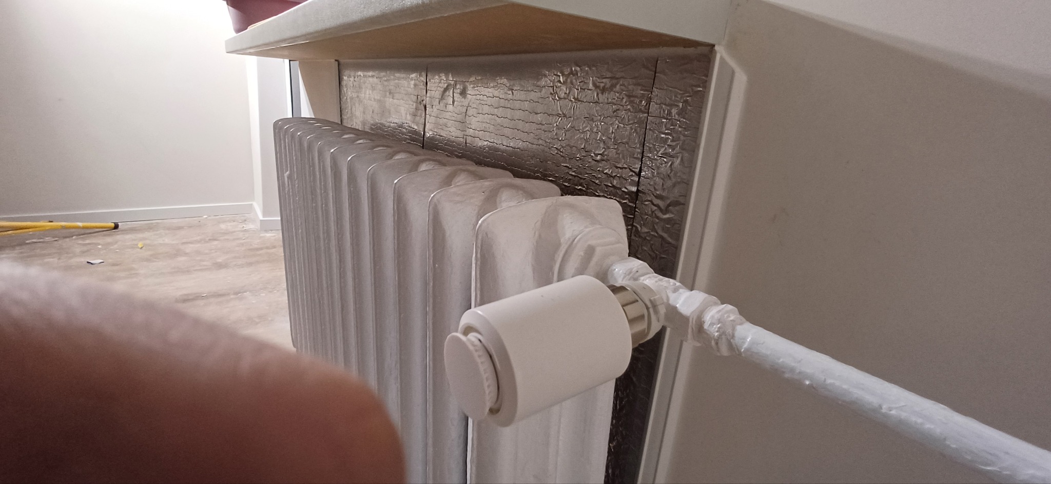 Biały, żeliwny grzejnik po renowacji, z nowym termostatem i zabezpieczonymi rurami, na tle ściany z izolacją termiczną. Widoczny fragment podłogi i drabina malarska.