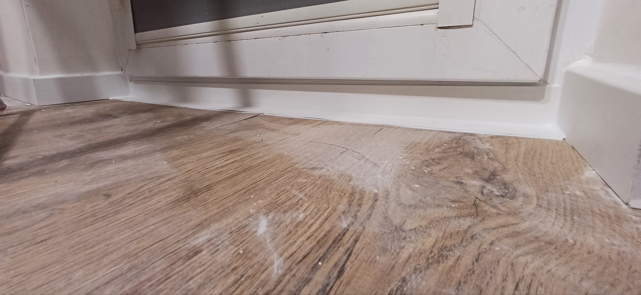 Białe listwy przypodłogowe połączone z panelami podłogowymi w odcieniach brązu. Widoczne ślady po remoncie. Ujęcie z niskiej perspektywy.