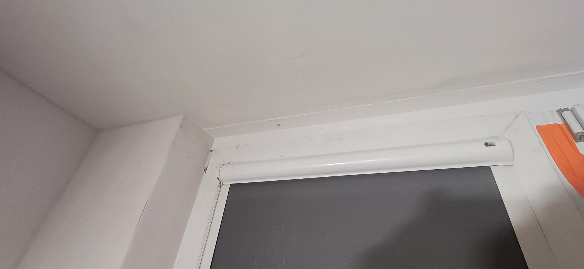 Biały sufit i ściana z widocznymi śladami po naprawach, narożnik i rama okienna z roletą. Widoczne niedoskonałości przed malowaniem.