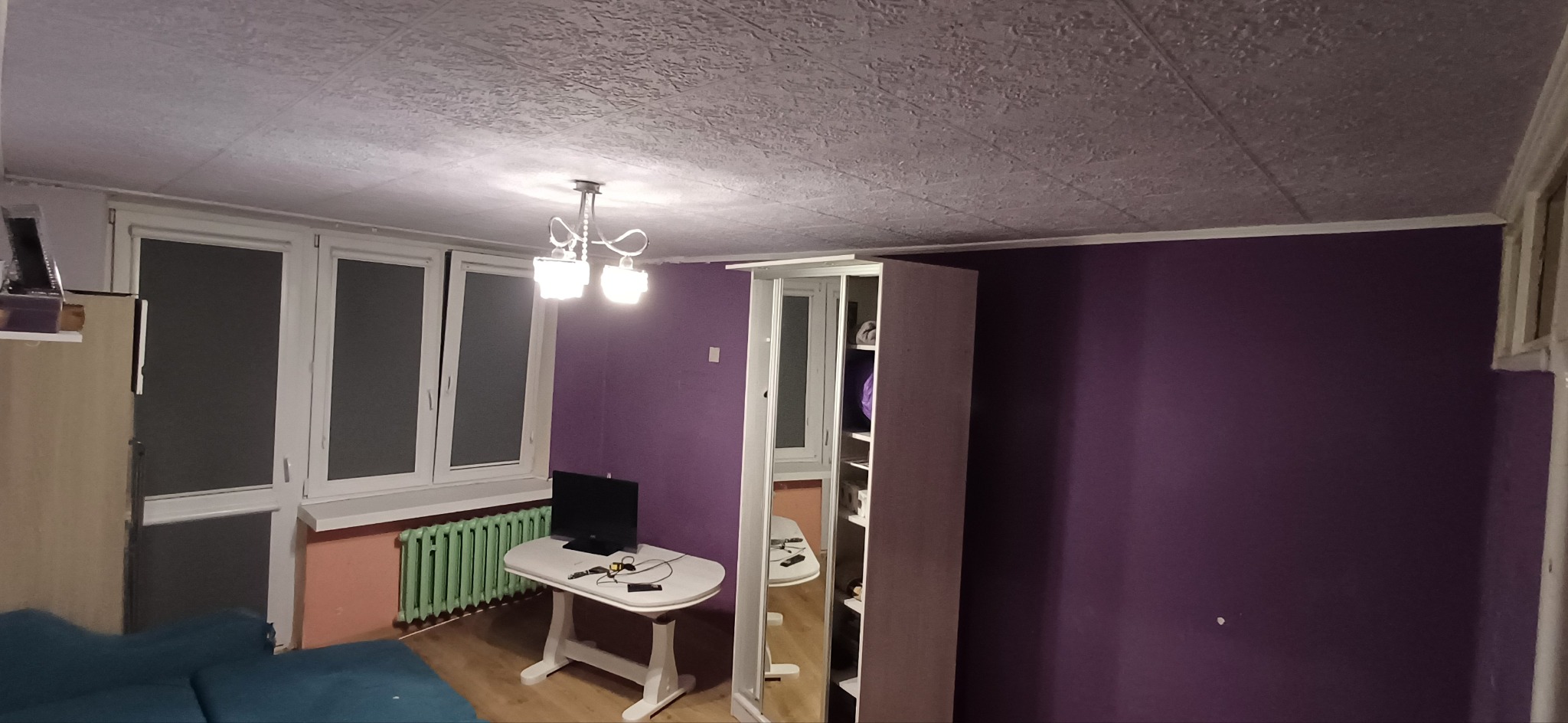 Fioletowa ściana w pokoju z oknami, grzejnikiem, stołem z telewizorem i szafą z lustrem. Sufit z fakturą. Widok na wnętrze po odświeżeniu.