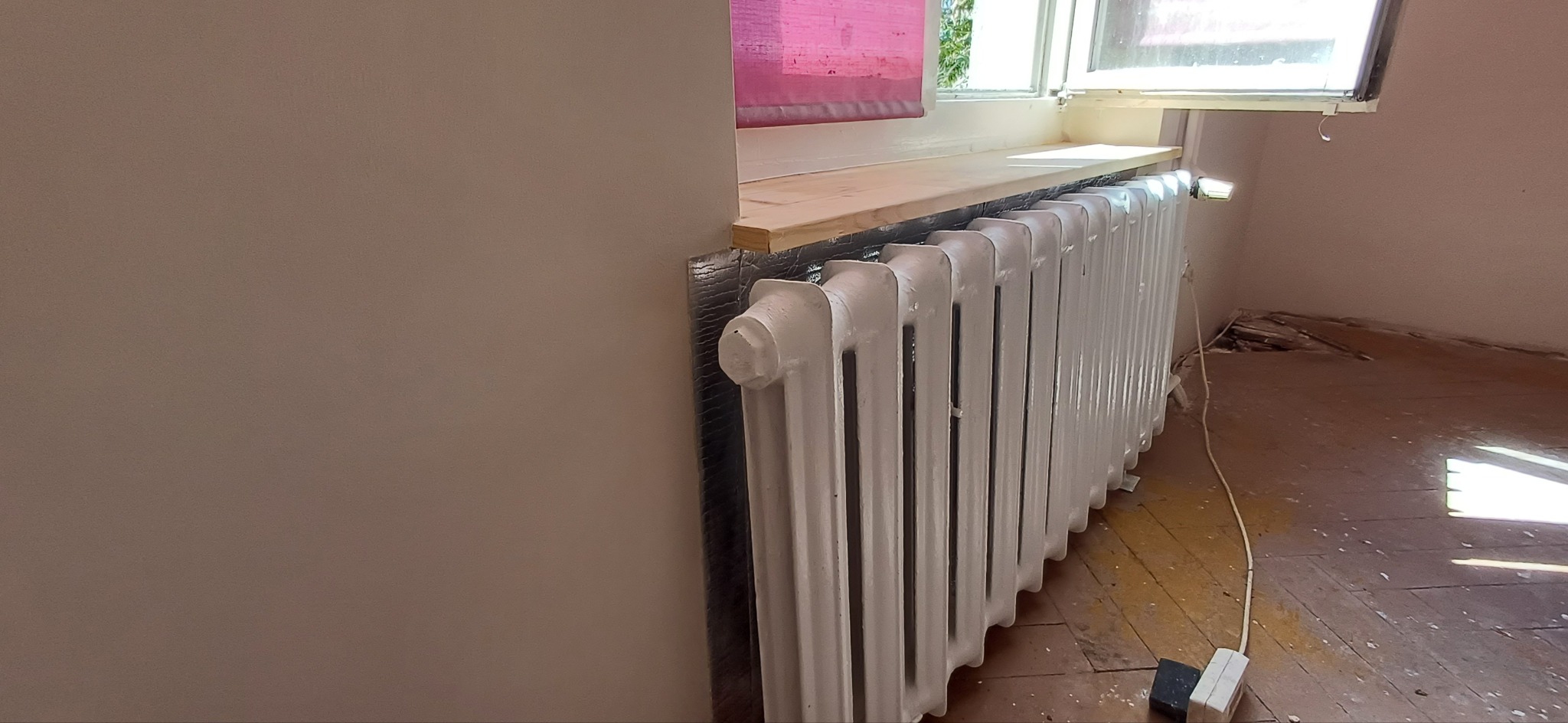 Biały, żeliwny grzejnik po renowacji, zamontowany pod oknem z drewnianym parapetem. Widoczne ślady prac remontowych na podłodze i świeżo pomalowana ściana.