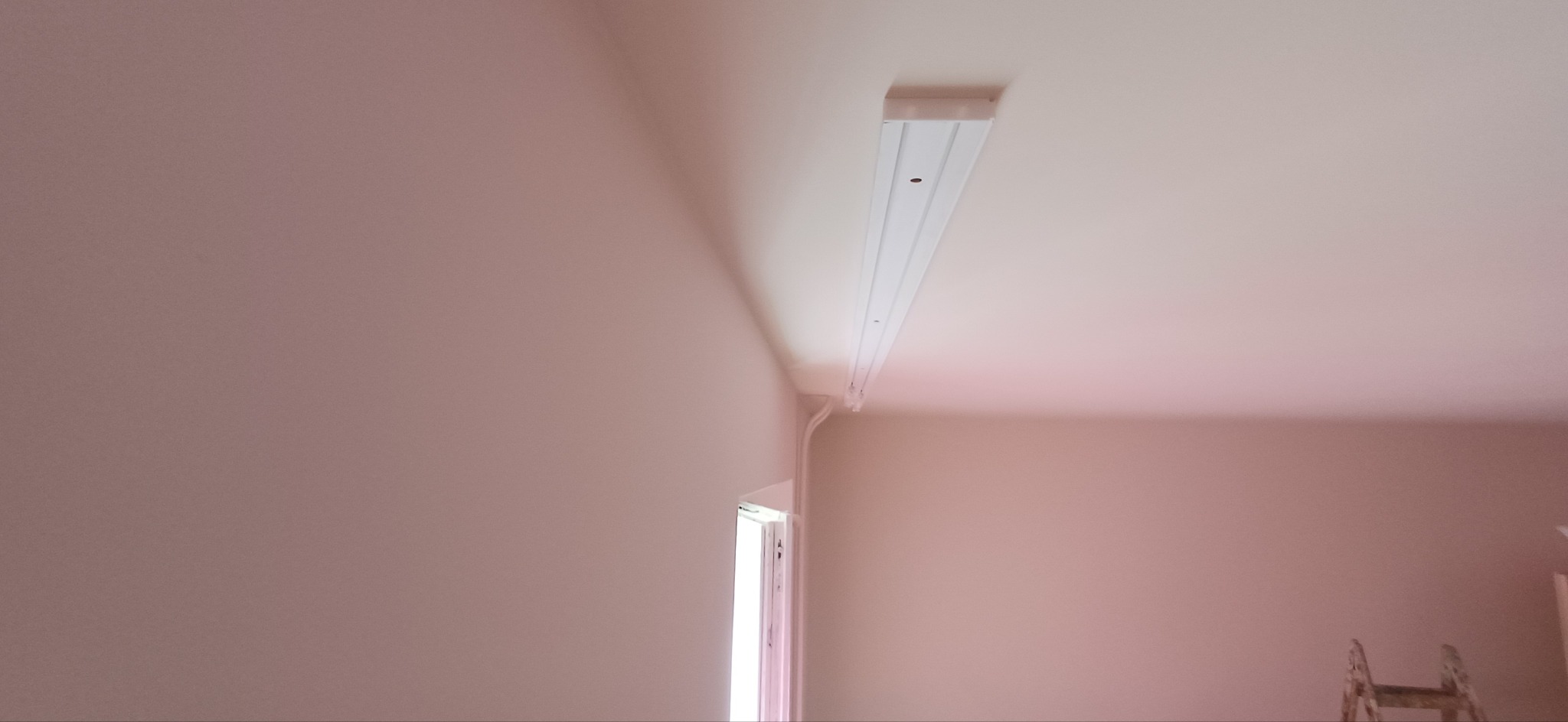 Zamontowana biała lampa sufitowa w pokoju o ścianach w odcieniach różu. Widoczny fragment okna i drabina. Ujęcie z dołu, podkreślające instalację oświetleniową.