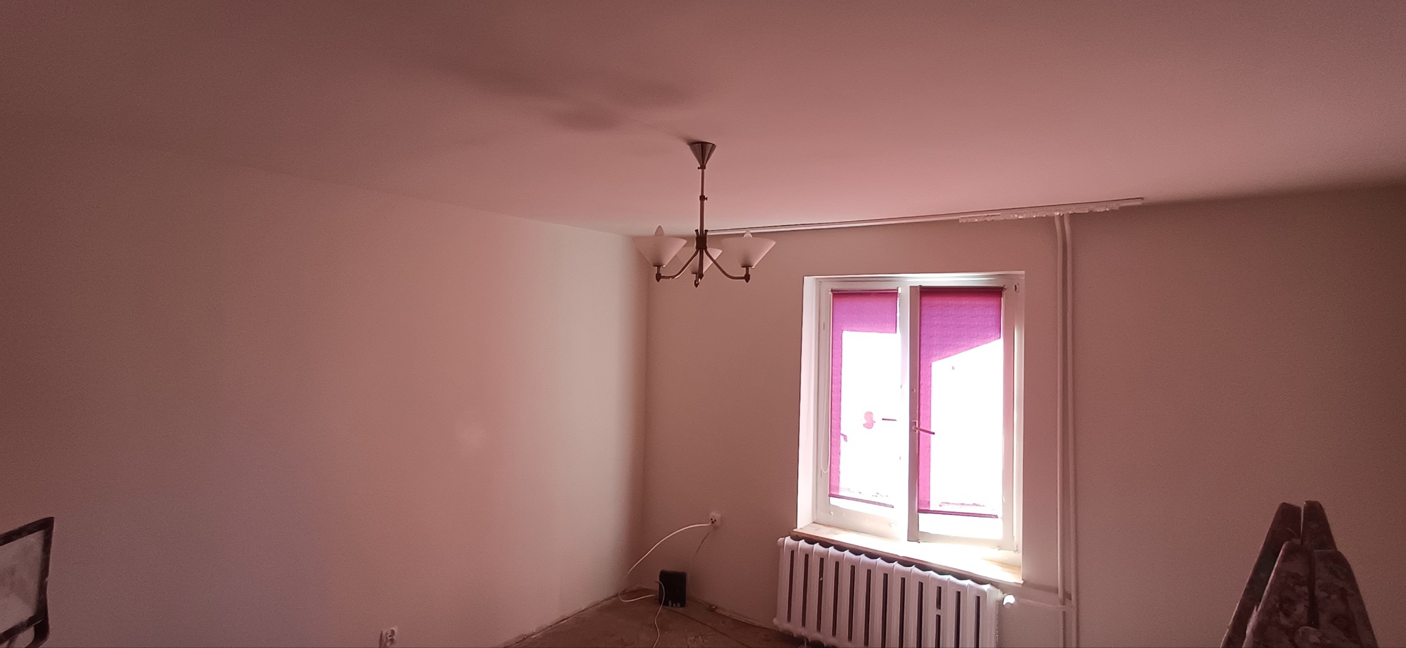 Jasne wnętrze po malowaniu ścian na gładko, z widocznym oknem z różowymi roletami i klasycznym żyrandolem. Ściany w odcieniach bieli i beżu.