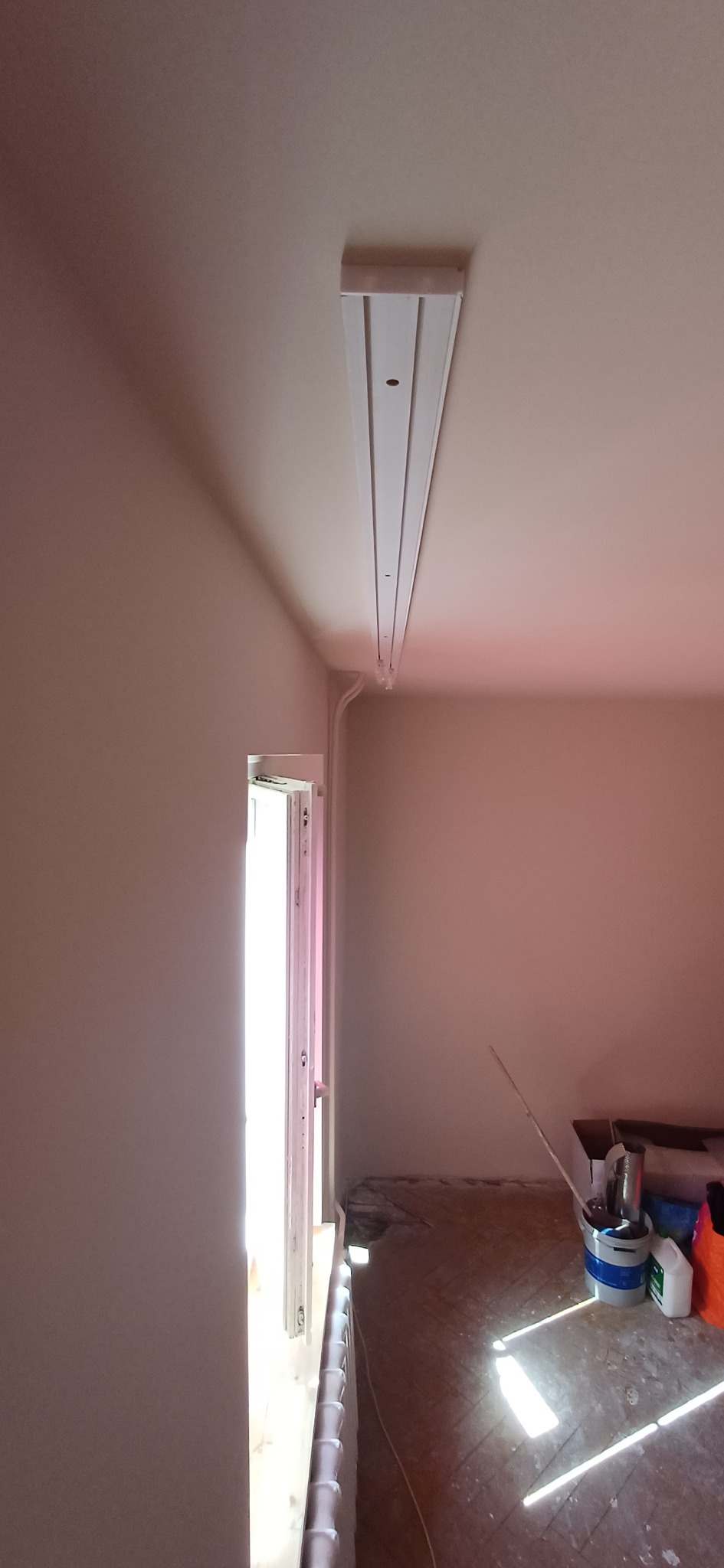 Wnętrze w trakcie remontu: pomalowane ściany na jasny kolor, zamontowana listwa oświetleniowa na suficie, widoczne narzędzia i materiały malarskie na podłodze.