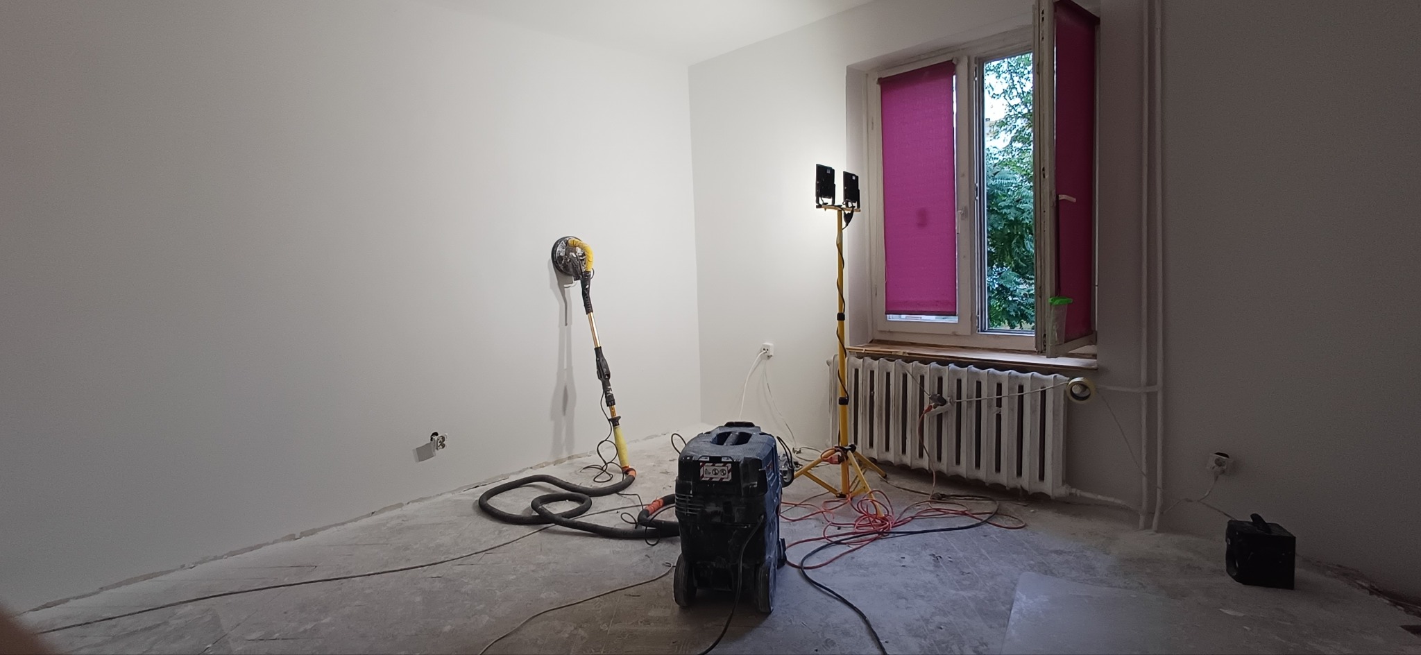 Pokój po szpachlowaniu ścian, widoczna szlifierka do gładzi, lampa oświetleniowa i odkurzacz przemysłowy. Ściany pomalowane na biało, okno z fioletowymi roletami.