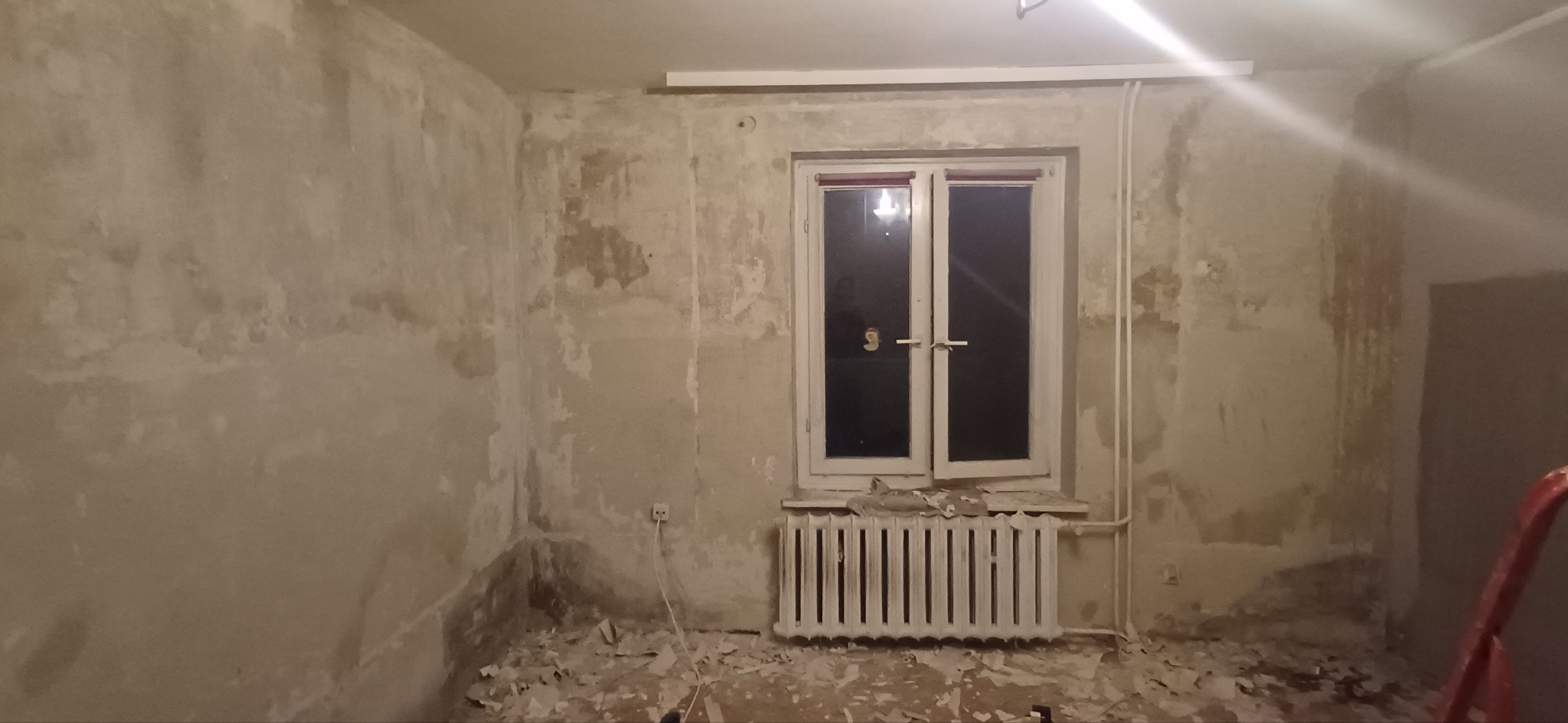 Pokój w trakcie przygotowań do remontu: ściany ze śladami starej farby, okno z grzejnikiem, resztki tapet na podłodze. Widoczne ślady po skuciu tynku.