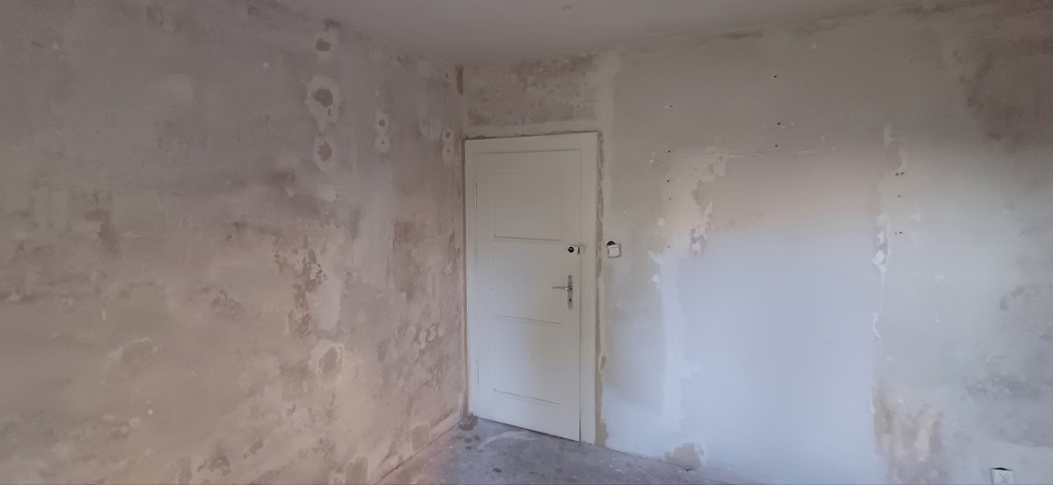Pokój w trakcie szpachlowania ścian, widoczne nierówności i ślady po łatach. Białe drzwi z klamką po prawej stronie. Przygotowanie do malowania.