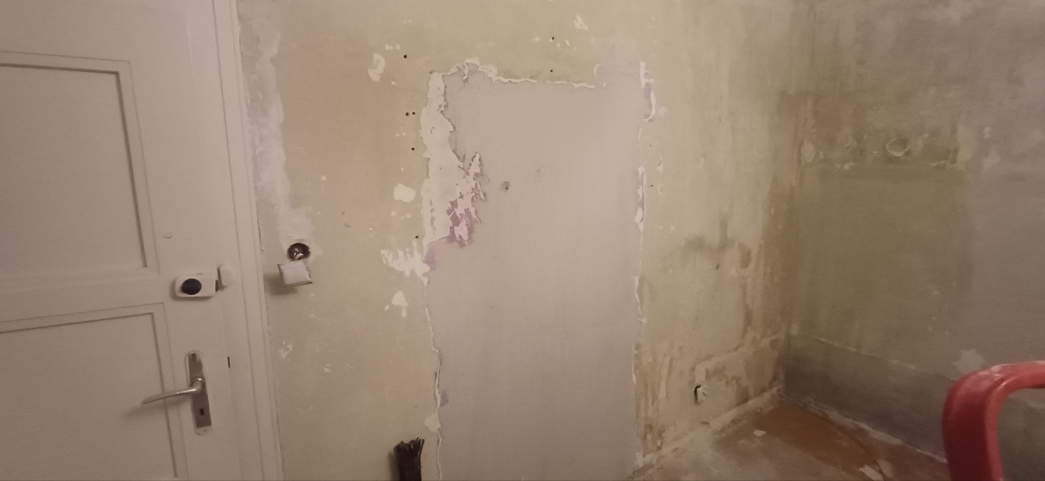 Ściana w trakcie przygotowania do malowania, widoczne ślady po usuniętej tapecie i gładzi, obok białe drzwi z metalową klamką. Fragment czerwonego krzesła w rogu.