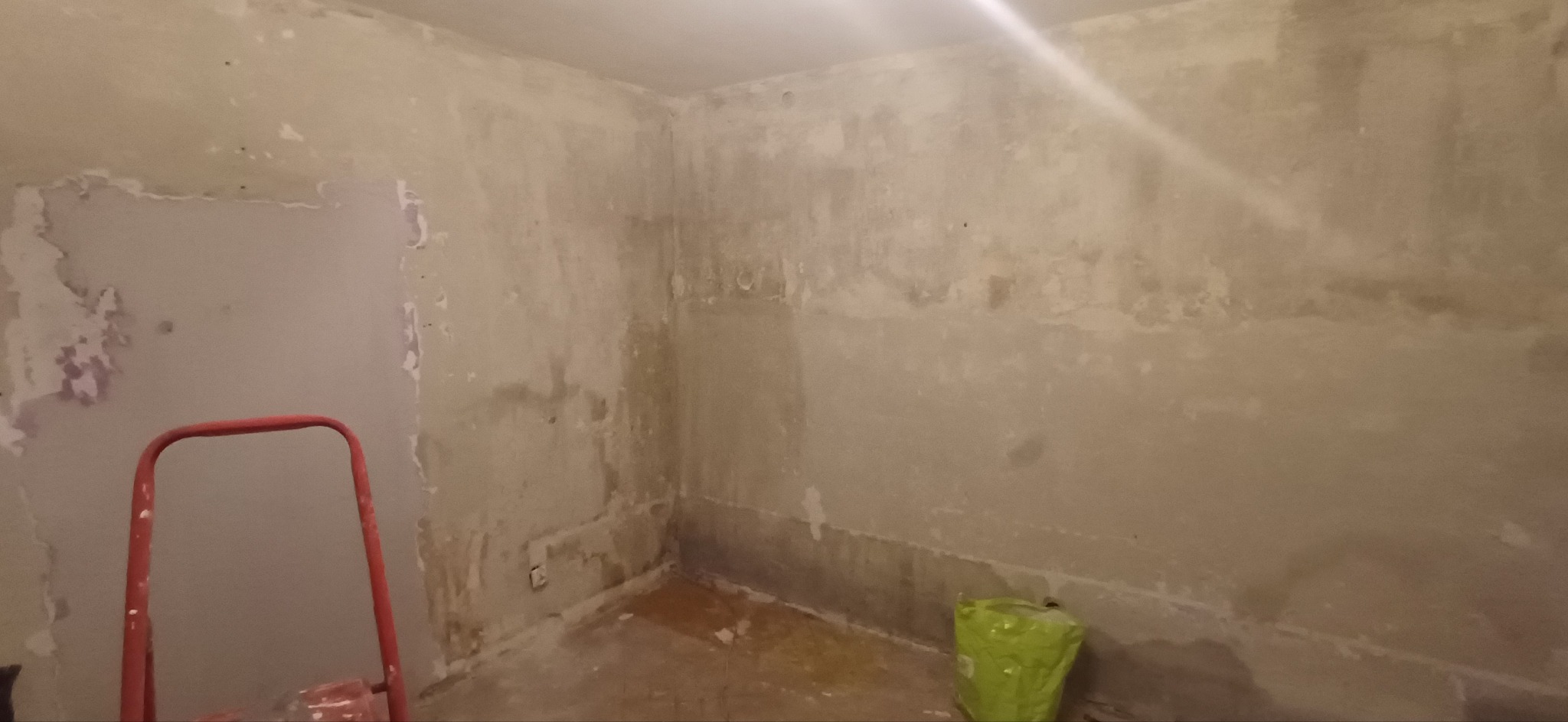 Pokój w trakcie przygotowania do malowania: ściany z resztkami starej farby, drabina malarska i worek z materiałami. Widoczne nierówności i ślady po usuniętych elementach.