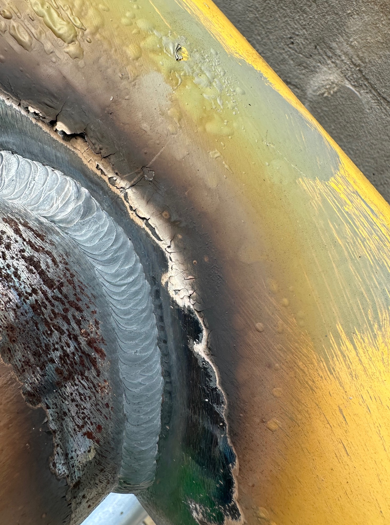 Zbliżenie na spoinę spawalniczą na żółtej, częściowo pomalowanej rurze. Widoczne ślady rdzy i odpryski farby, ukazujące precyzję i trwałość wykonanego złącza.