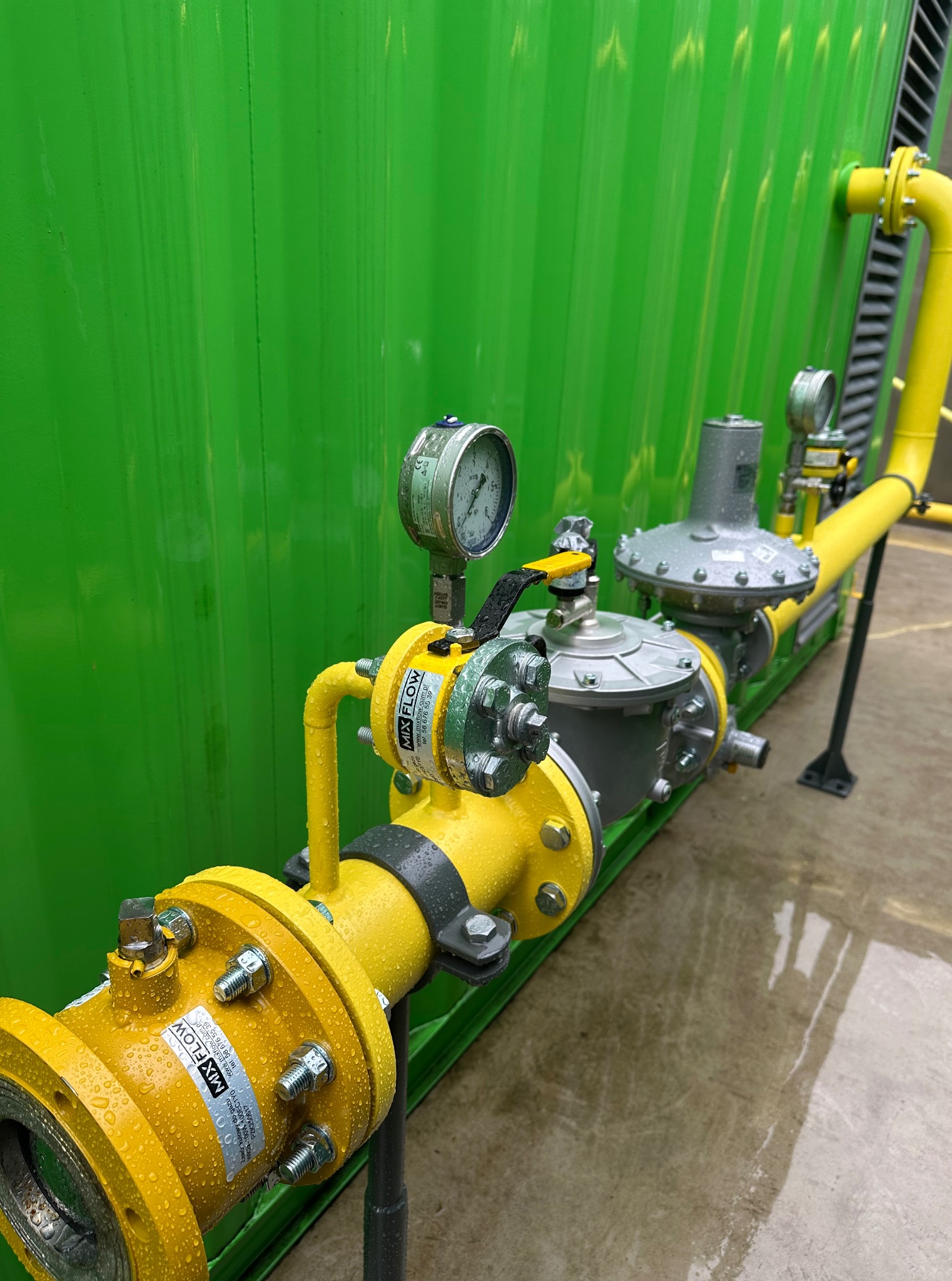 Żółta instalacja gazowa z manometrem, złączkami kołnierzowymi i zaworami, zamontowana na tle zielonej ściany kontenera. Widoczne krople wody.