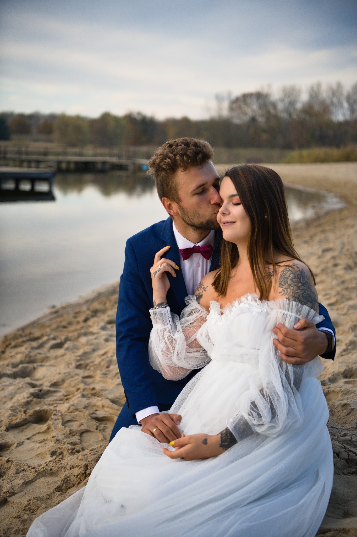 Para młoda na plaży: on w granatowym garniturze z bordową muchą, całuje pannę młodą w białej sukni z tatuażami na ramionach. W tle woda i drewniany pomost.