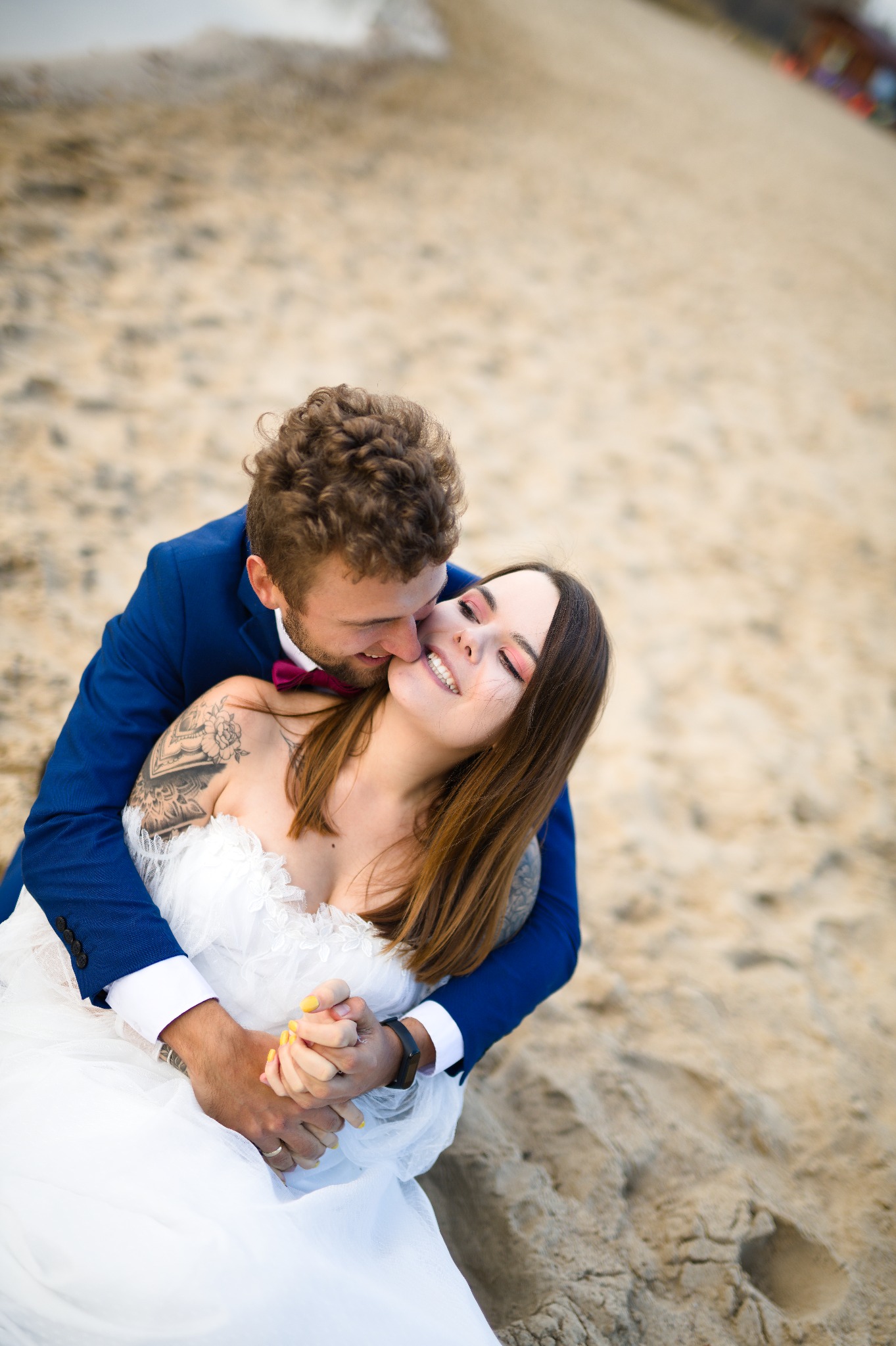 Uśmiechnięta panna młoda w objęciach pana młodego na plaży. Ona w białej sukni, on w niebieskim garniturze z bordową muchą. Widoczne tatuaże na ramieniu panny młodej.