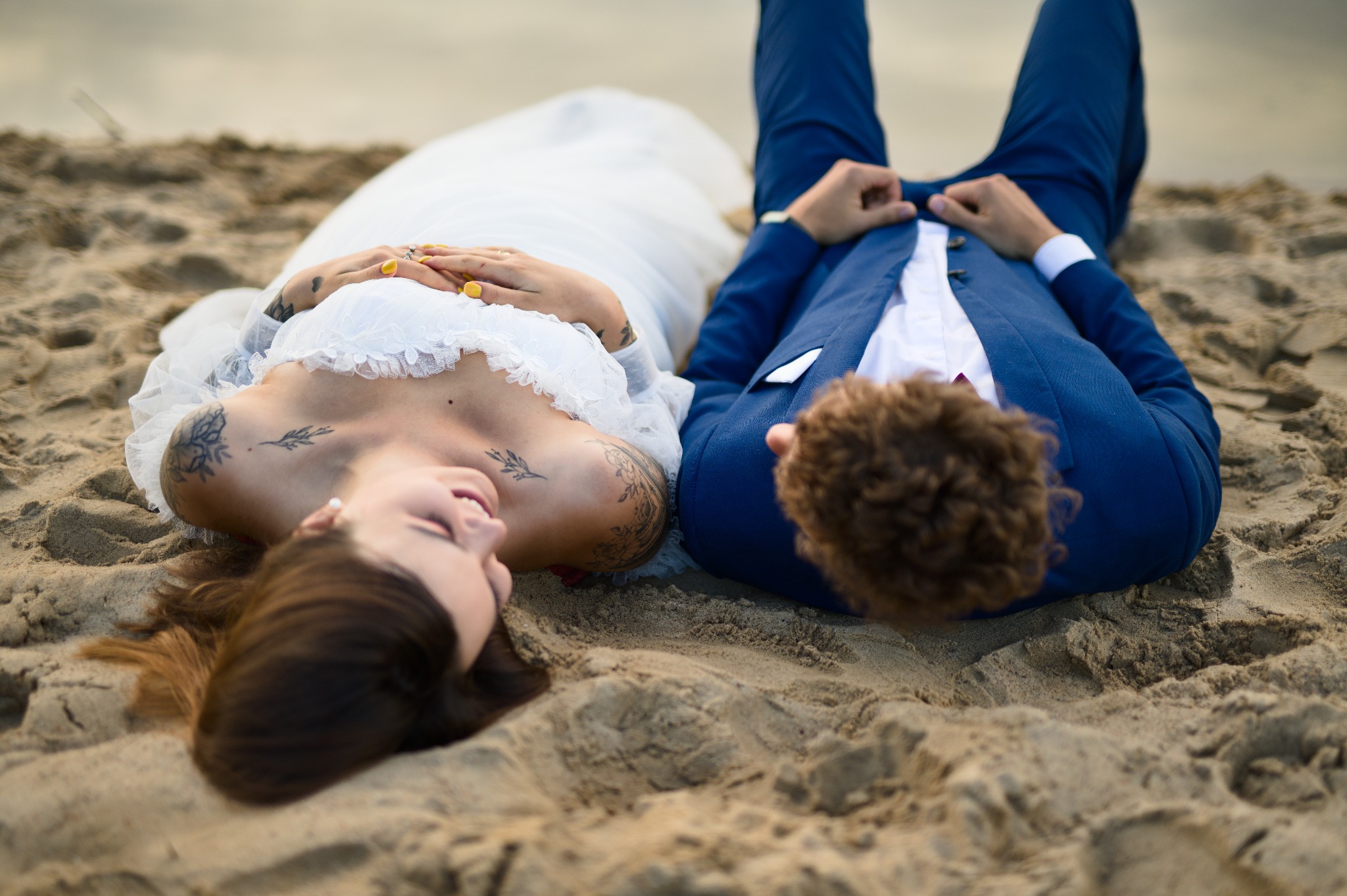 Para młoda leżąca na piasku w zbliżeniu: panna młoda w białej sukni z tatuażami i pan młody w niebieskim garniturze. Romantyczna sesja ślubna na plaży.
