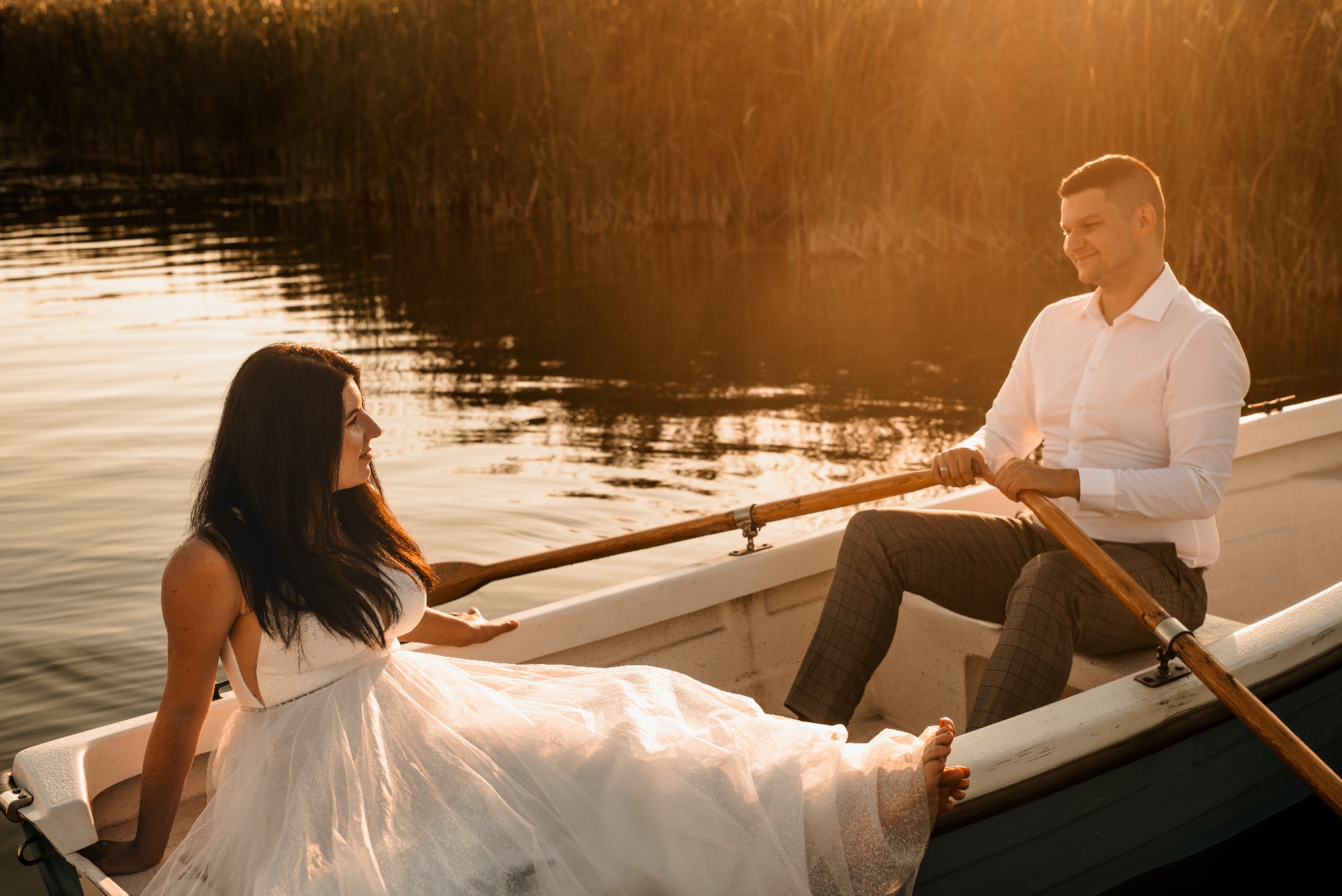 Sesja ślubna w Bydgoszczy: Para młoda w łódce na jeziorze o zachodzie słońca. Panna młoda w białej sukni, pan młody wiosłuje. Romantyczna atmosfera.