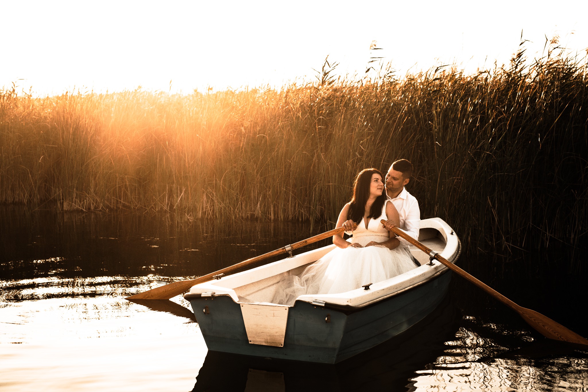 Para młoda w łódce na tle trzcin oświetlonych zachodzącym słońcem. Romantyczna sesja ślubna na wodzie, delikatne światło, naturalne otoczenie.