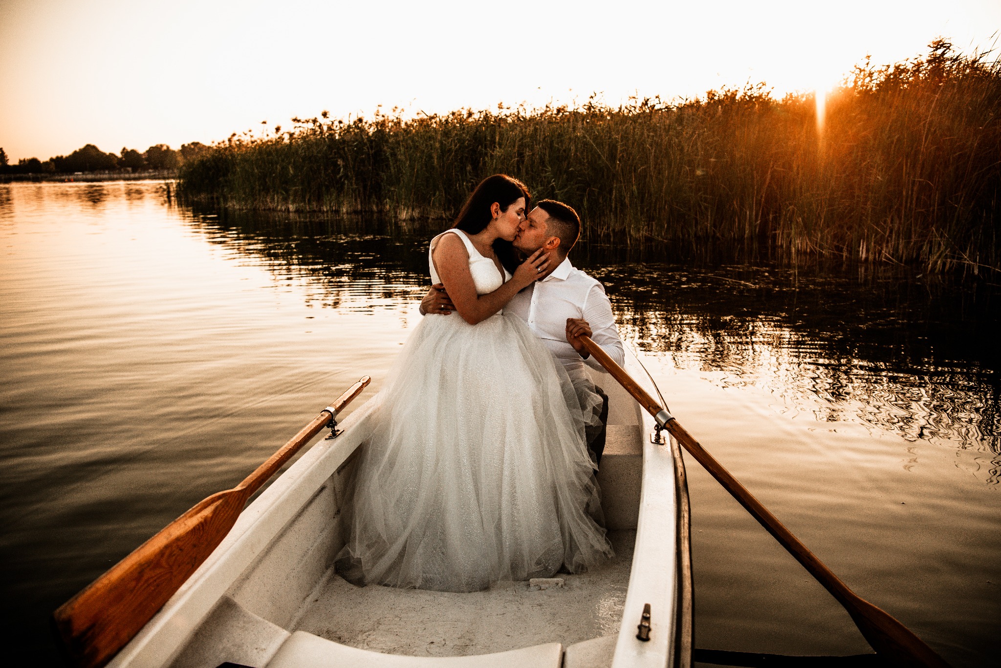 Pocałunek pary młodej w łódce na tle trzcin i zachodzącego słońca. Romantyczna sesja plenerowa z wiosłami i wodą. Ciepłe, złote światło.