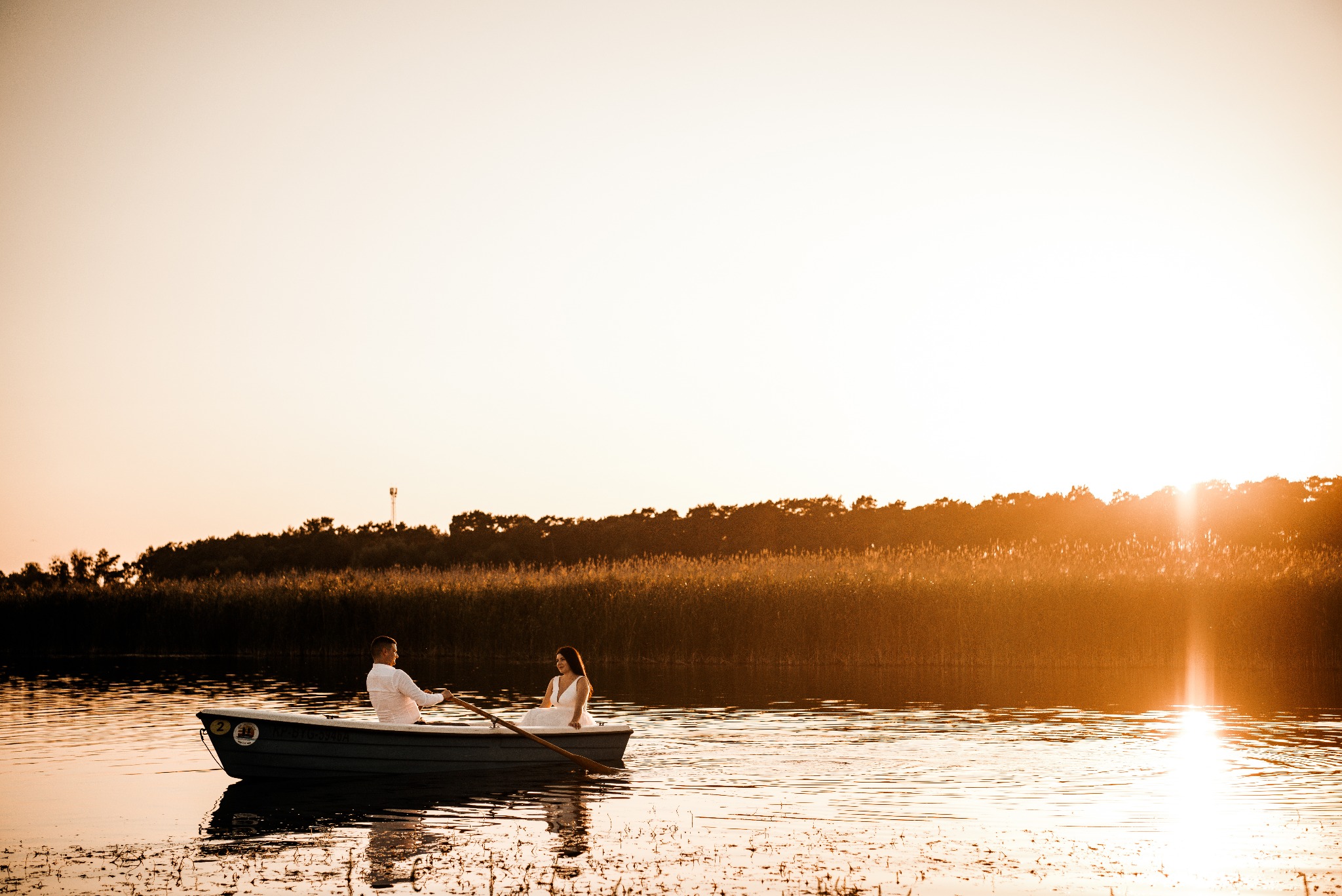 Para młoda w łódce na jeziorze o zachodzie słońca. Pan młody wiosłuje, panna młoda siedzi obok. Romantyczna sceneria z promieniami słońca odbijającymi się w wodzie.