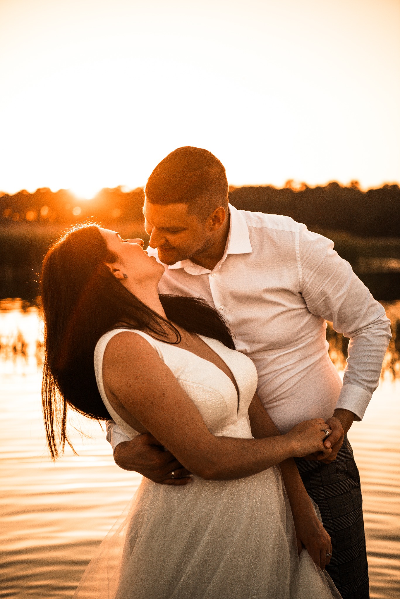 Para młoda w objęciach na tle jeziora o zachodzie słońca. Panna młoda w białej sukni, pan młody w białej koszuli i spodniach w kratę. Romantyczna sesja ślubna.