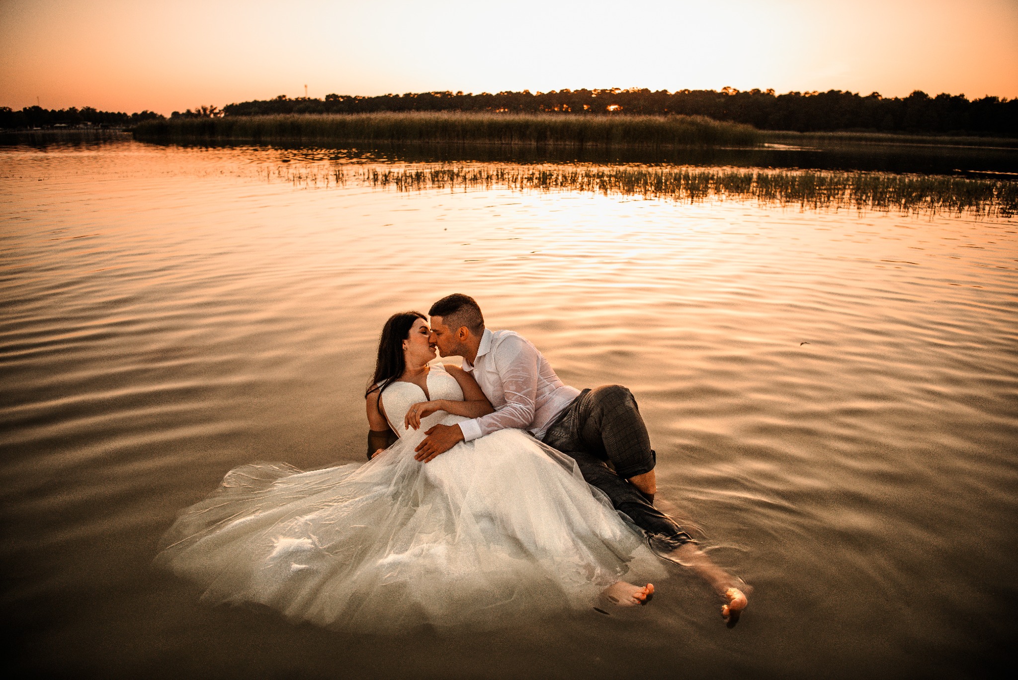 Para młoda w wodzie podczas zachodu słońca. Panna młoda w białej sukni, pan młody w mokrej koszuli. Romantyczna sesja ślubna na tle jeziora i lasu.