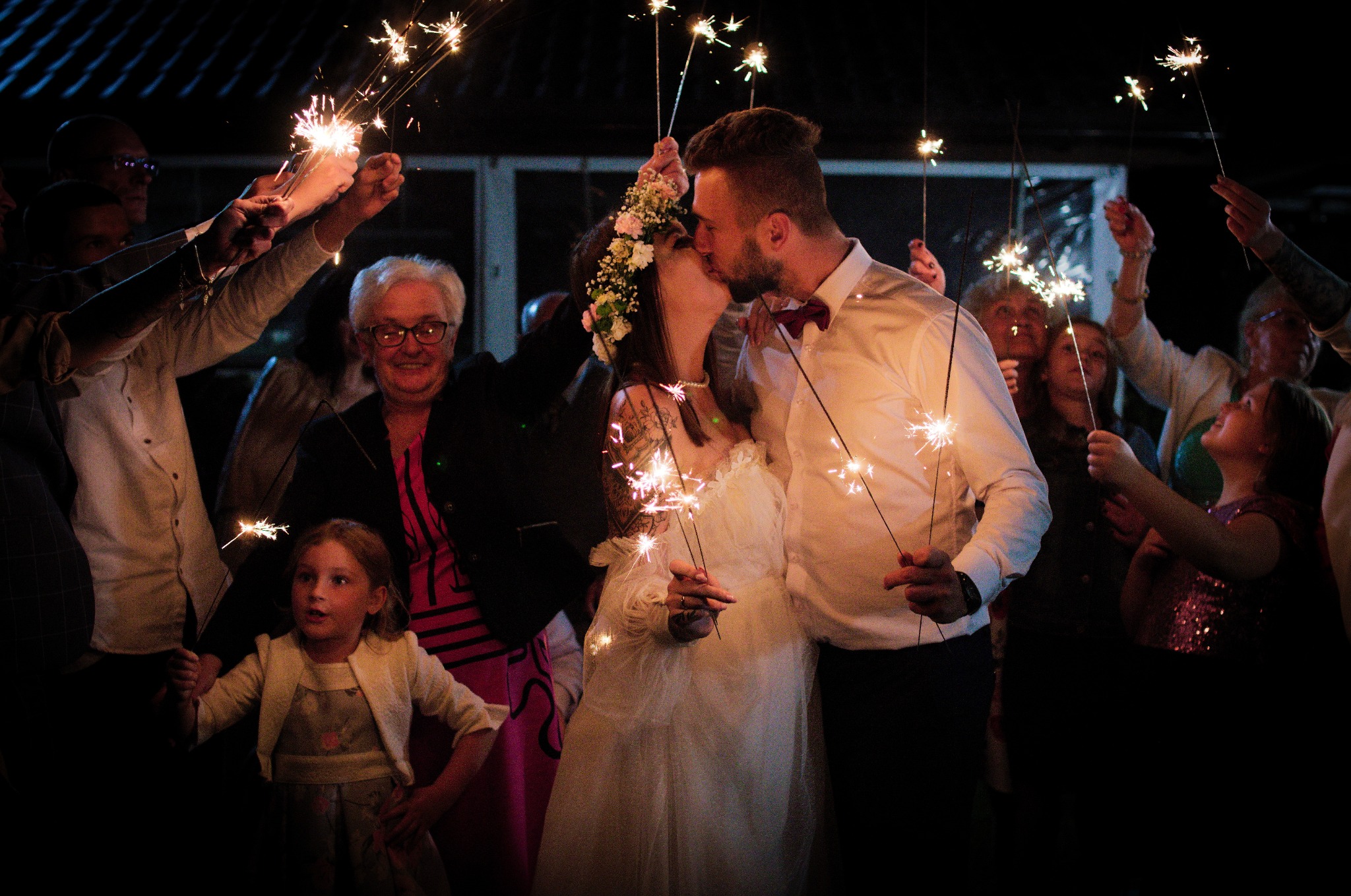 Pocałunek pary młodej otoczonej gośćmi z zimnymi ogniami. Panna młoda w wianku i sukni, pan młody w muszce. Uśmiechnięte twarze w ciemnym otoczeniu.