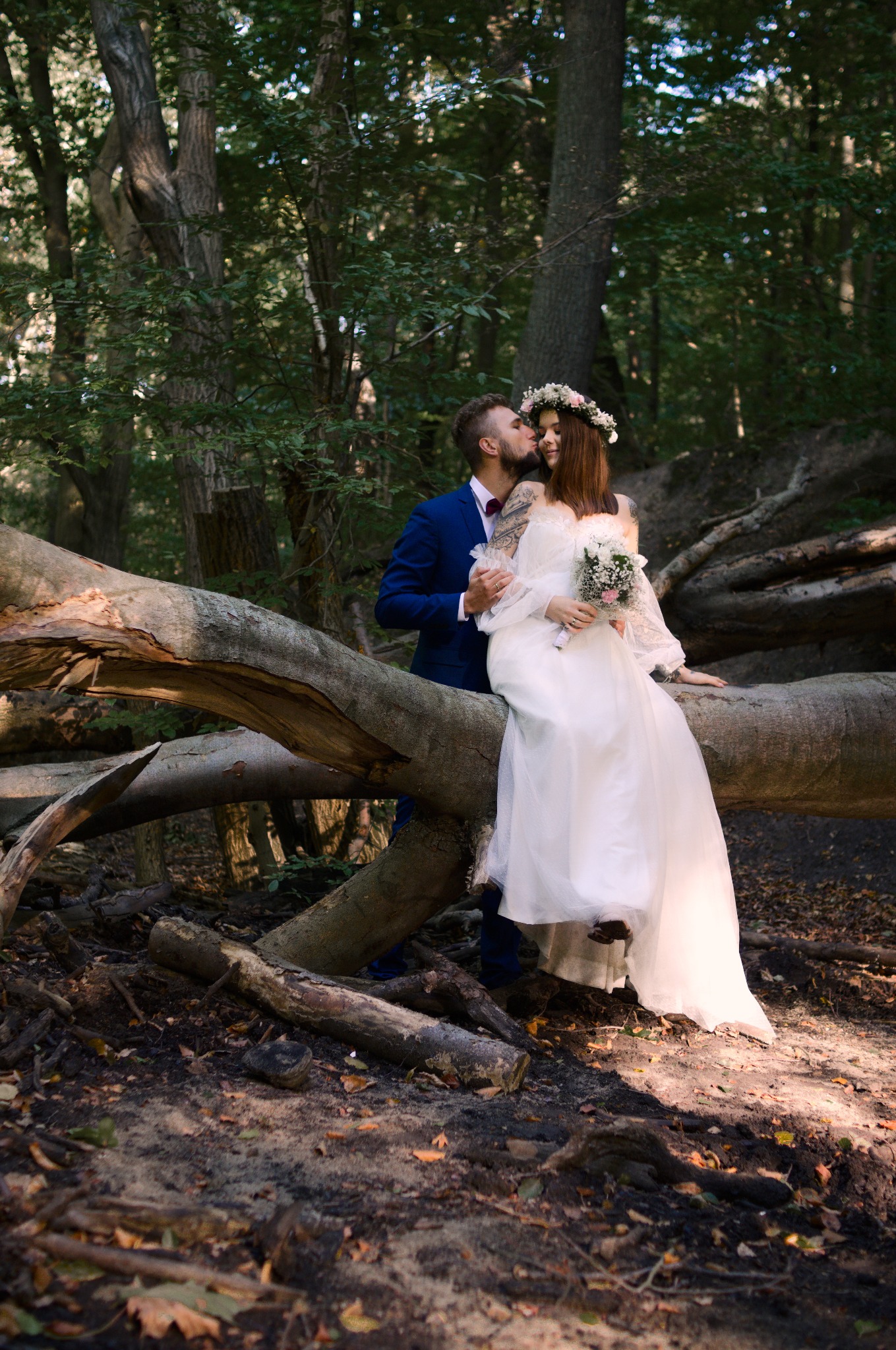 Para młoda w leśnej scenerii: panna młoda w wianku i sukni siedzi na powalonym drzewie, pan młody w niebieskim garniturze całuje ją w policzek. Romantyczna sesja ślubna.