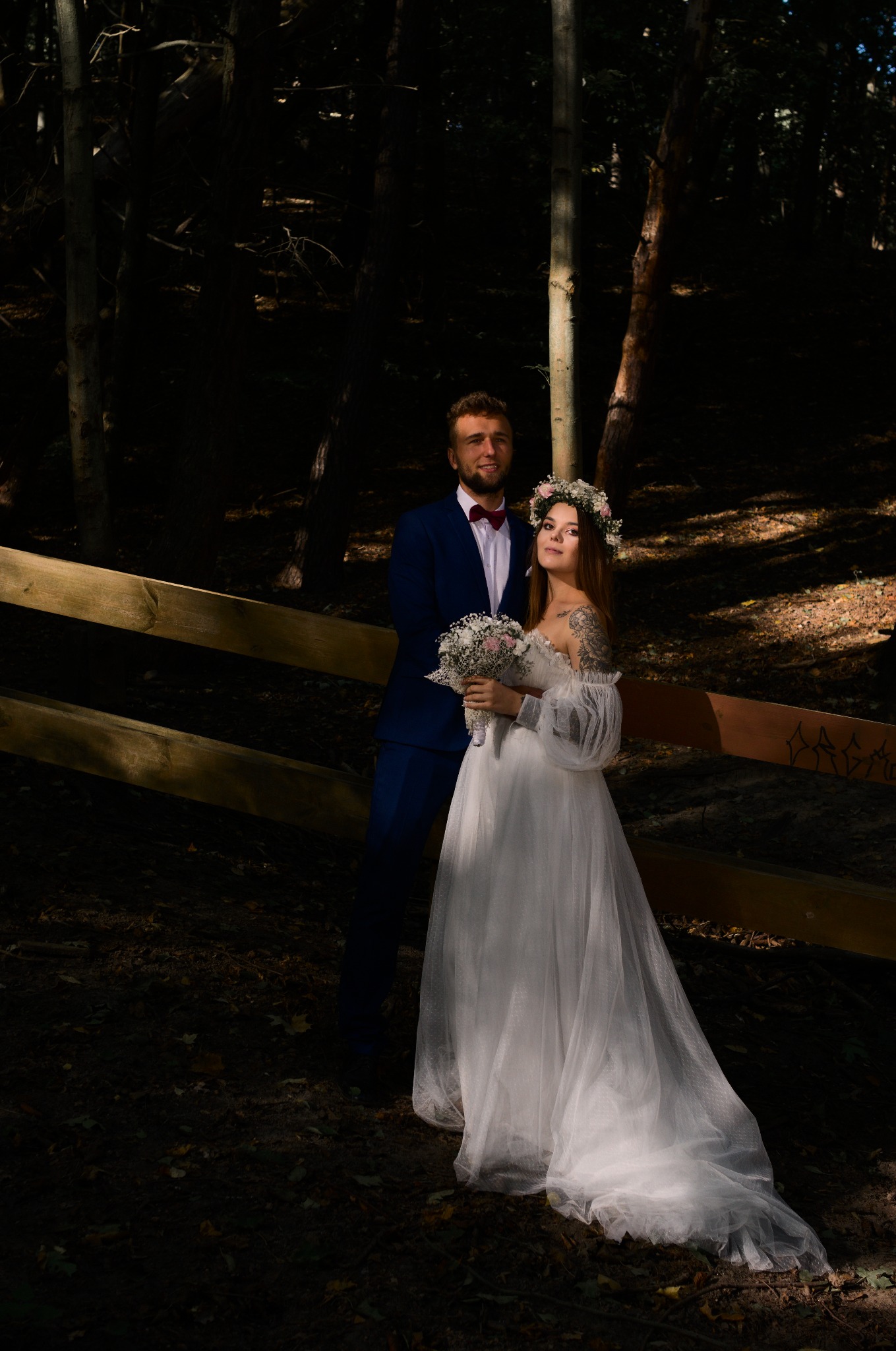 Para młoda w lesie: panna młoda w wianku i sukni z bufiastymi rękawami, pan młody w granatowym garniturze i bordowej muszce, opierają się o drewniane ogrodzenie.