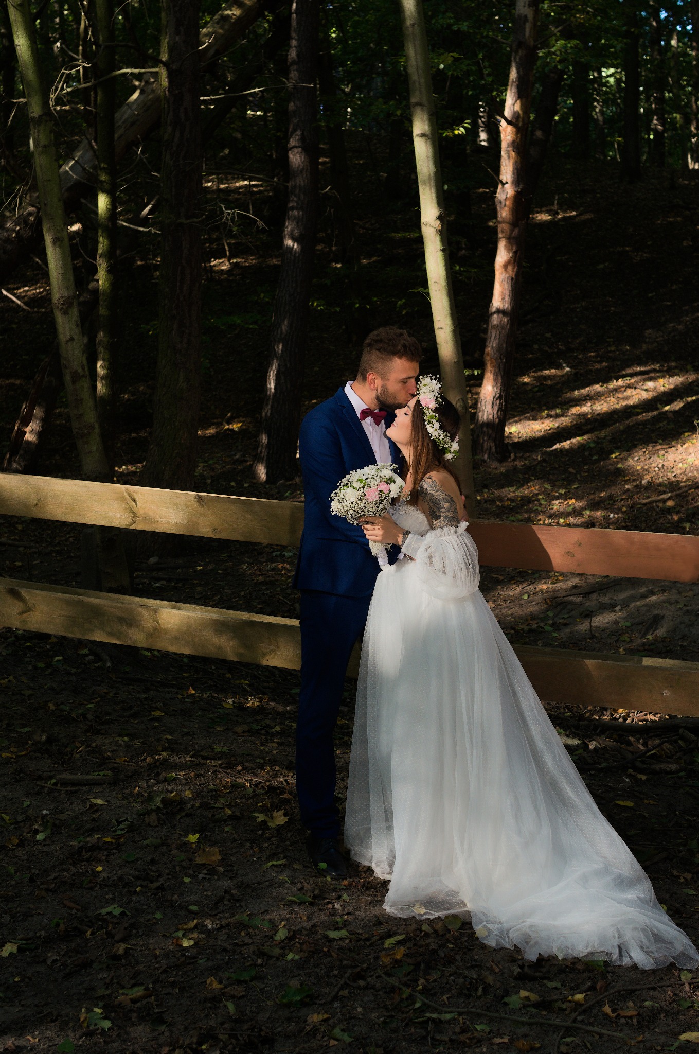 Para młoda w leśnej scenerii: pan młody w granatowym garniturze całuje pannę młodą w wianku i zwiewnej sukni, trzymającą bukiet. Romantyczna sesja przy drewnianym płocie.