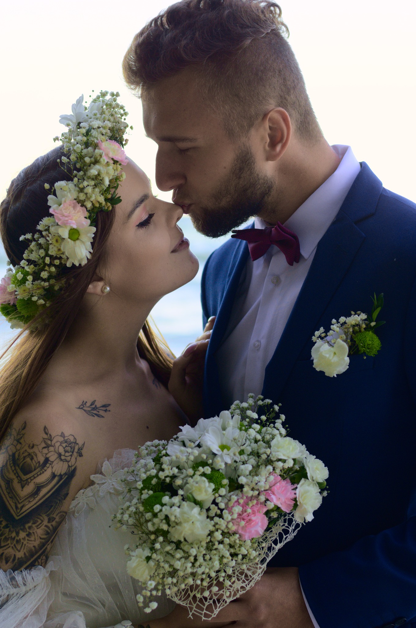 Pocałunek młodej pary: panna młoda w wianku i sukni z koronką, z tatuażem na ramieniu, trzyma bukiet; pan młody w granatowym garniturze z bordową muchą.