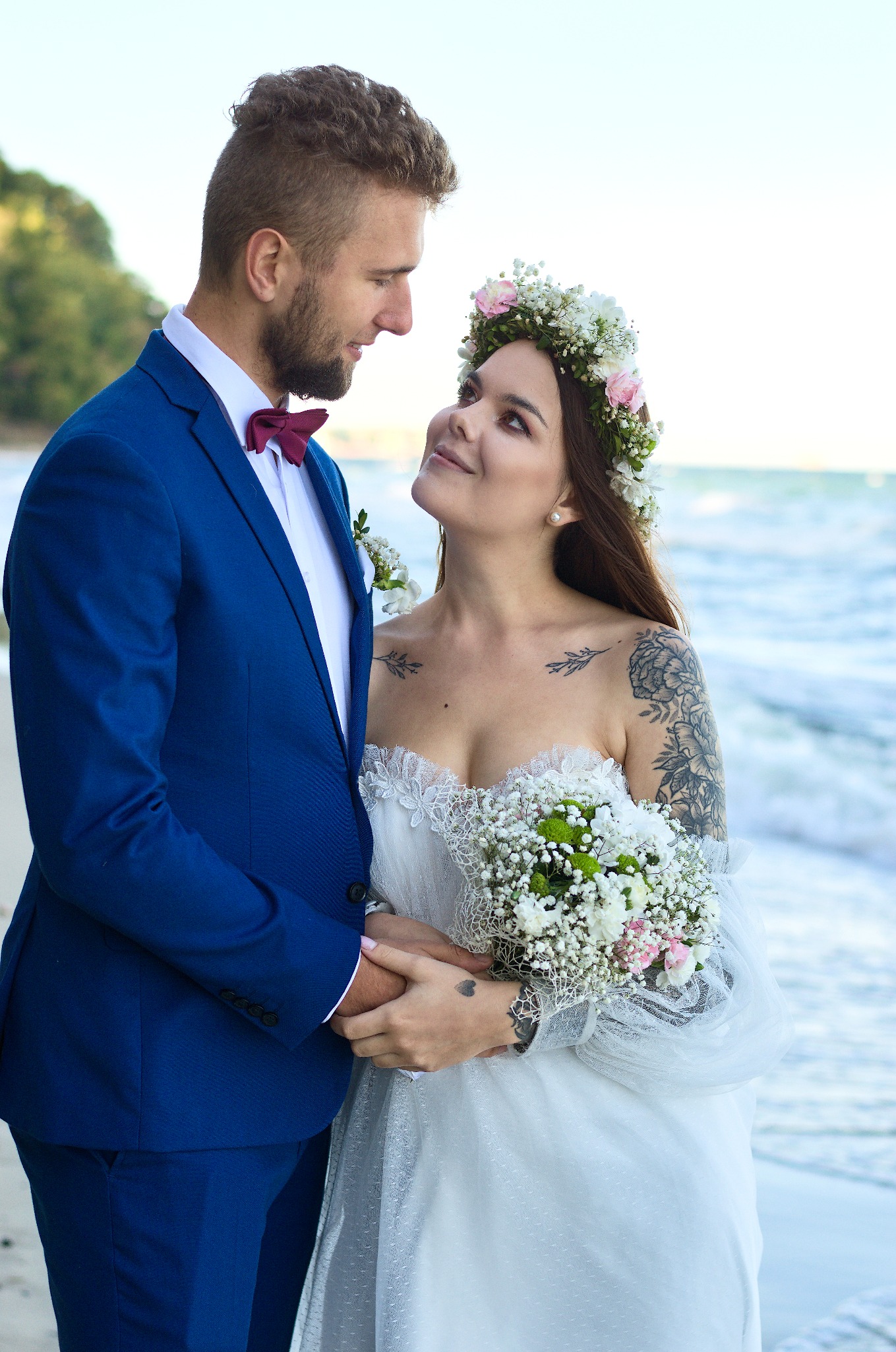 Para młoda na plaży: on w granatowym garniturze, ona w białej sukni z wiankiem, trzymają się za ręce, patrząc na siebie z czułością. W tle morskie fale i zieleń.