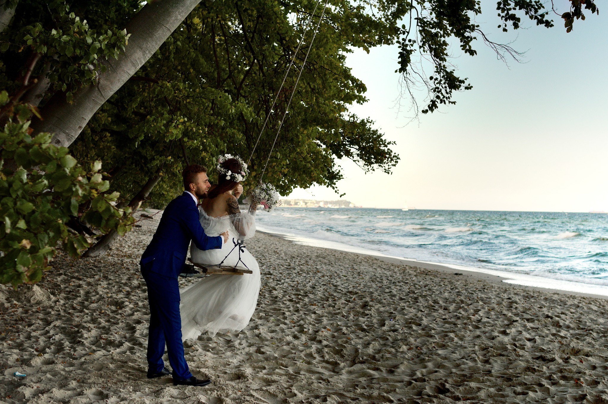 Para młoda na huśtawce na plaży, on w granatowym garniturze, ona w białej sukni z wiankiem. W tle morze i drzewa. Romantyczna sesja ślubna w plenerze.