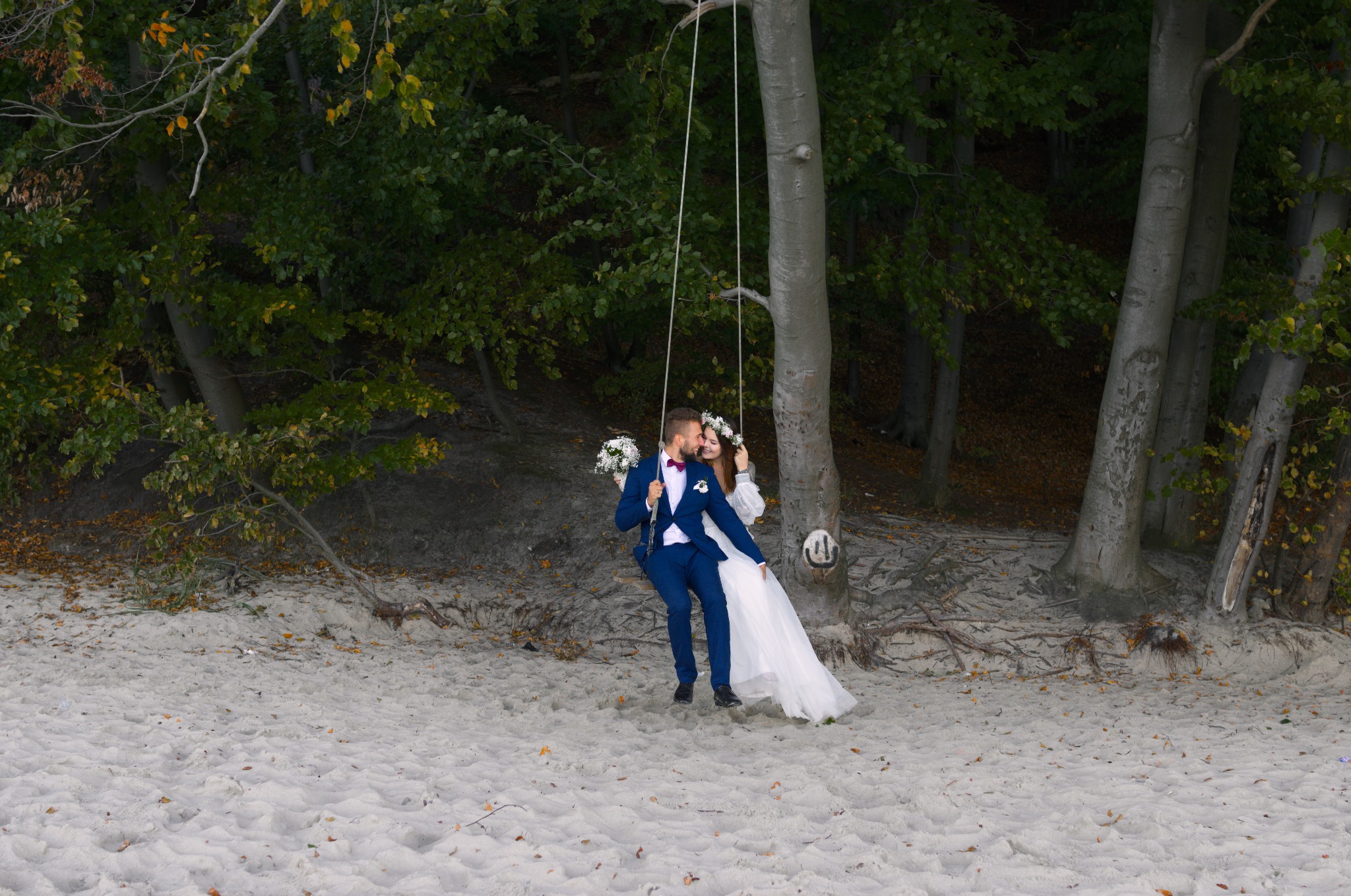 Para młoda w strojach ślubnych siedzi na huśtawce zawieszonej na drzewie na piaszczystej plaży, otoczona zielenią lasu. Romantyczna sesja w plenerze.