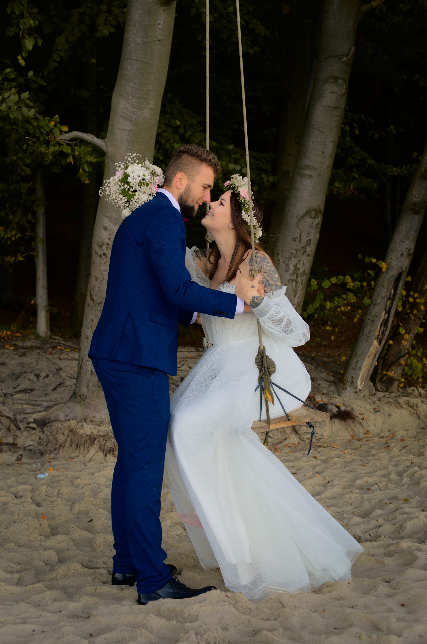 Para młoda na huśtawce z desek na plaży. Panna młoda w koronie z kwiatów i sukni z koronkowymi rękawami, pan młody w granatowym garniturze. Romantyczna sesja ślubna.
