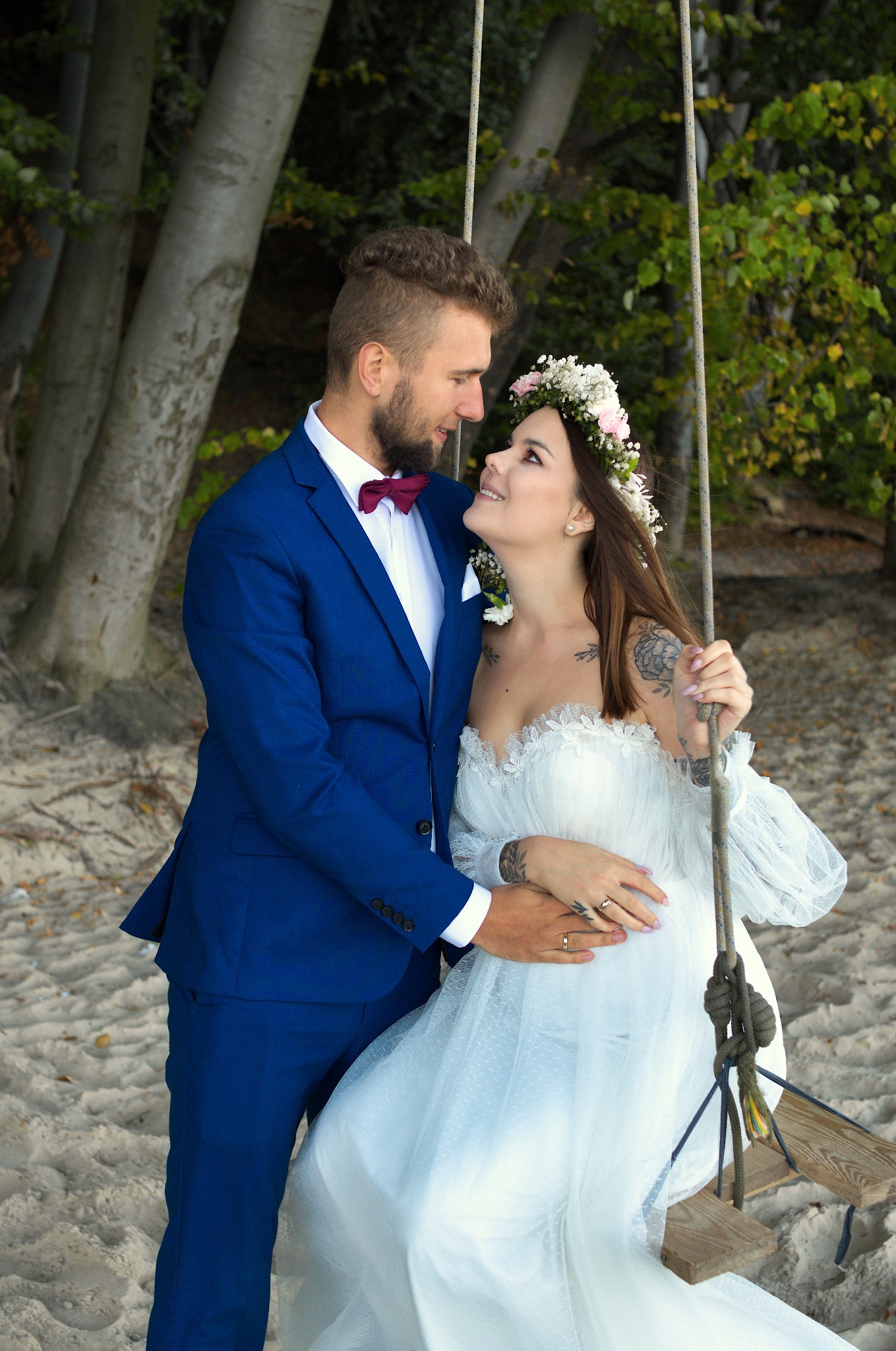 Uśmiechnięta para młoda na huśtawce na plaży. Pan młody w niebieskim garniturze z bordową muchą, panna młoda w białej sukni z wiankiem. Romantyczna sesja w plenerze.