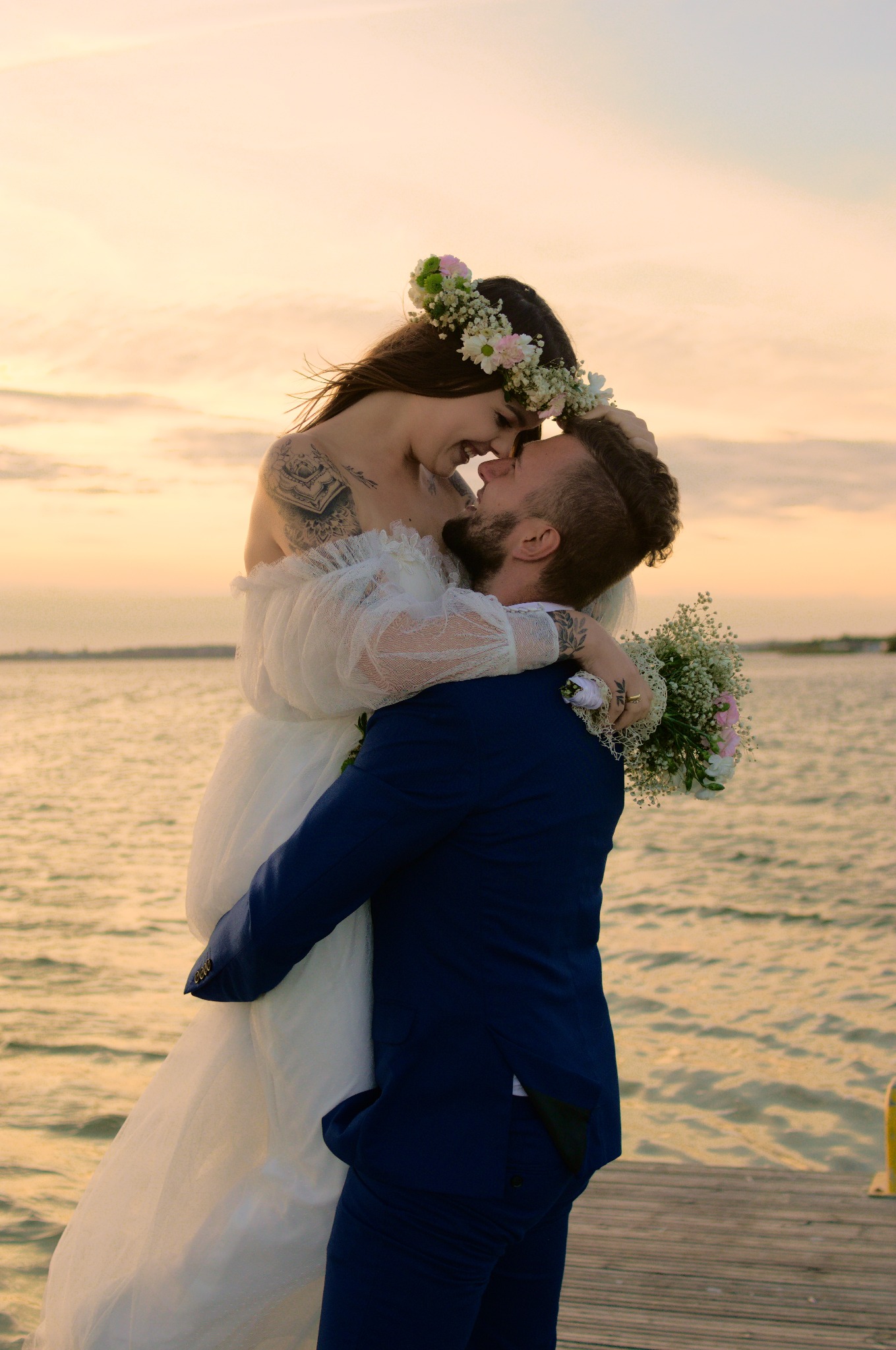 Para młoda na pomoście: panna młoda w wianku kwiatów, z tatuażem na ramieniu, uniesiona przez pana młodego w granatowym garniturze na tle zachodzącego słońca nad wodą.
