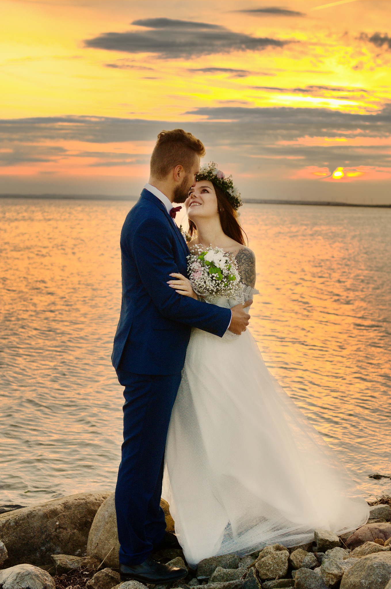 Para młoda w objęciach na tle zachodzącego słońca nad wodą. Panna młoda w wianku i z tatuażem na ramieniu, pan młody w granatowym garniturze.