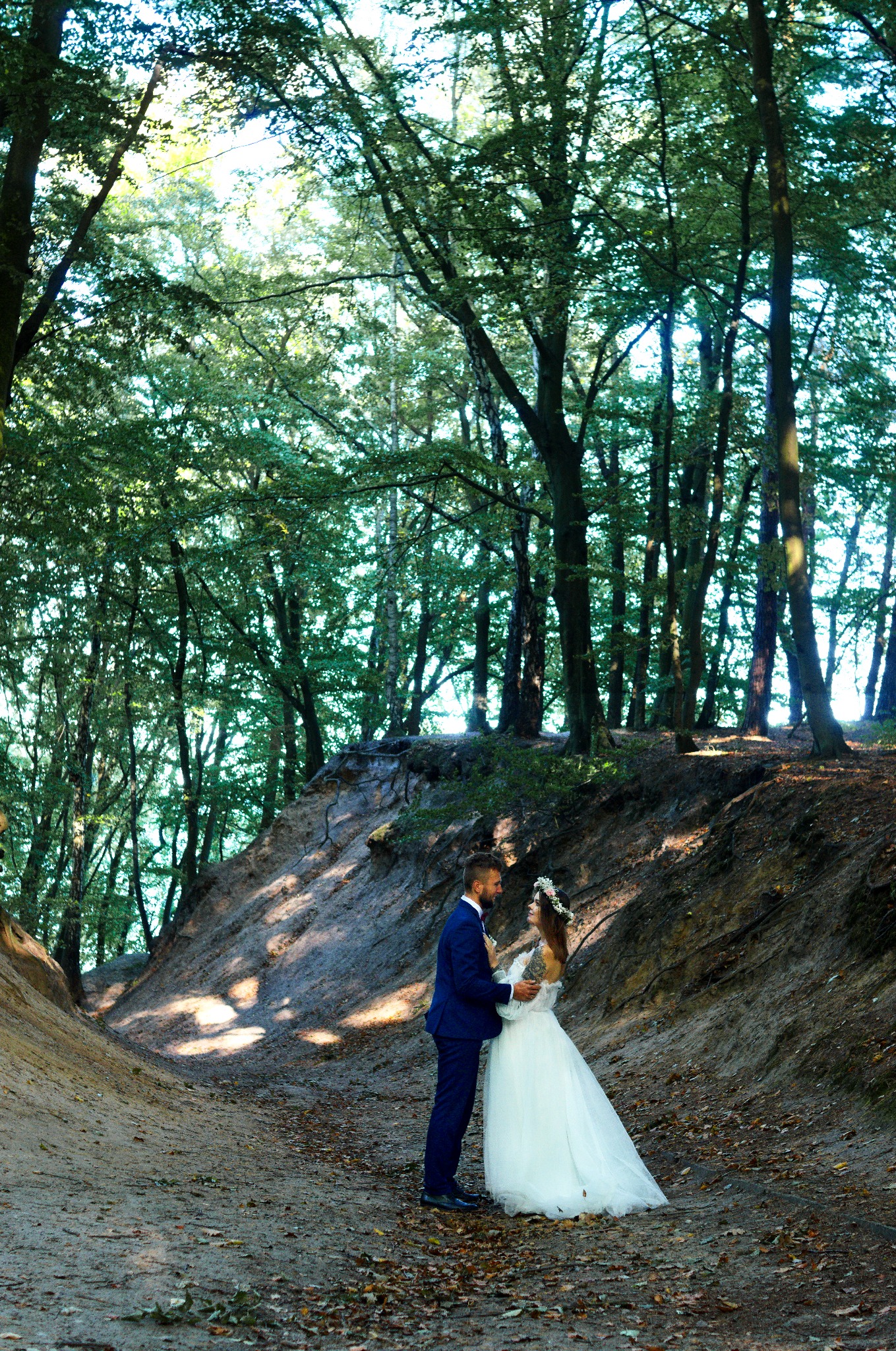 Para młoda w leśnej scenerii: panna młoda w białej sukni i wianku, pan młody w granatowym garniturze, drzewa w tle, naturalne światło, romantyczna atmosfera.