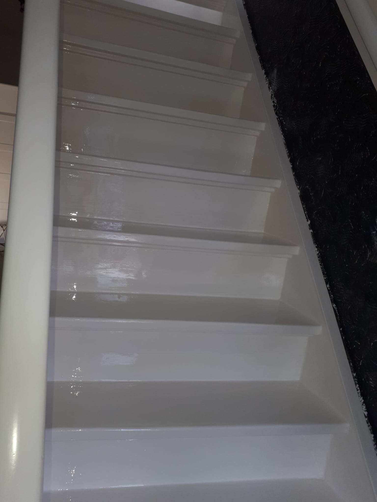 Białe, świeżo pomalowane schody wewnętrzne z widoczną strukturą farby i ciemnym tłem ściany po prawej stronie. Ujęcie z dołu, podkreślające głębię perspektywy.