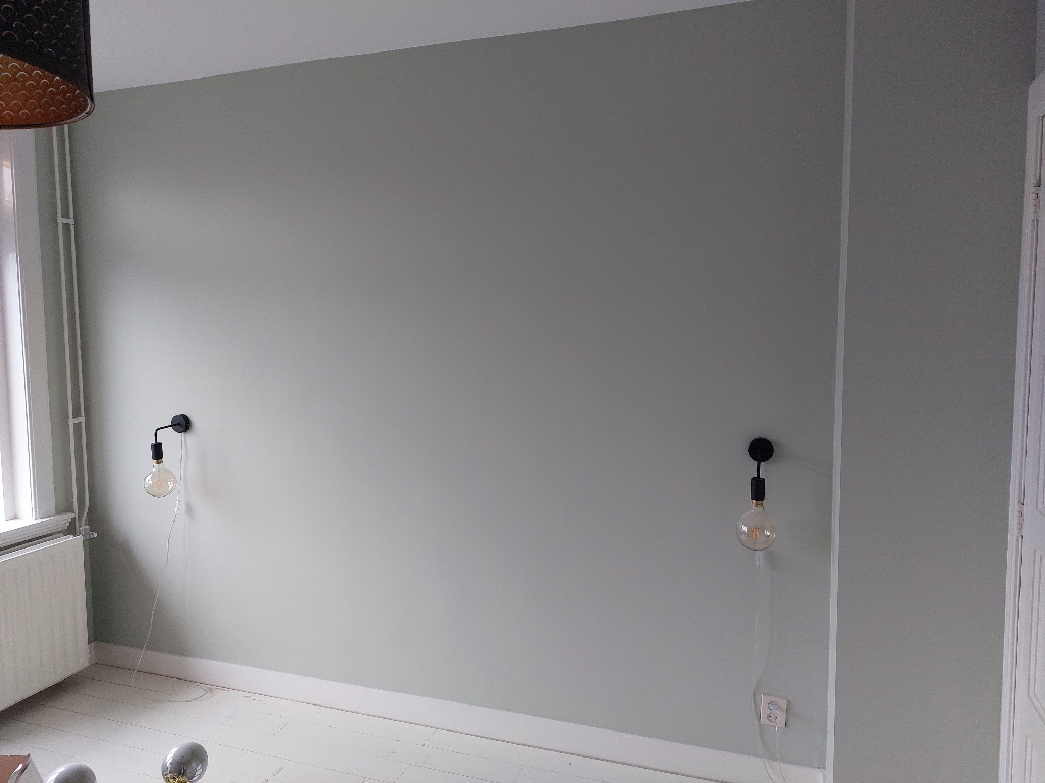 Ściana w odcieniach szarości po odświeżeniu, z dwoma lampami ściennymi z żarówkami Edisona. Widoczne białe listwy przypodłogowe i fragment grzejnika.
