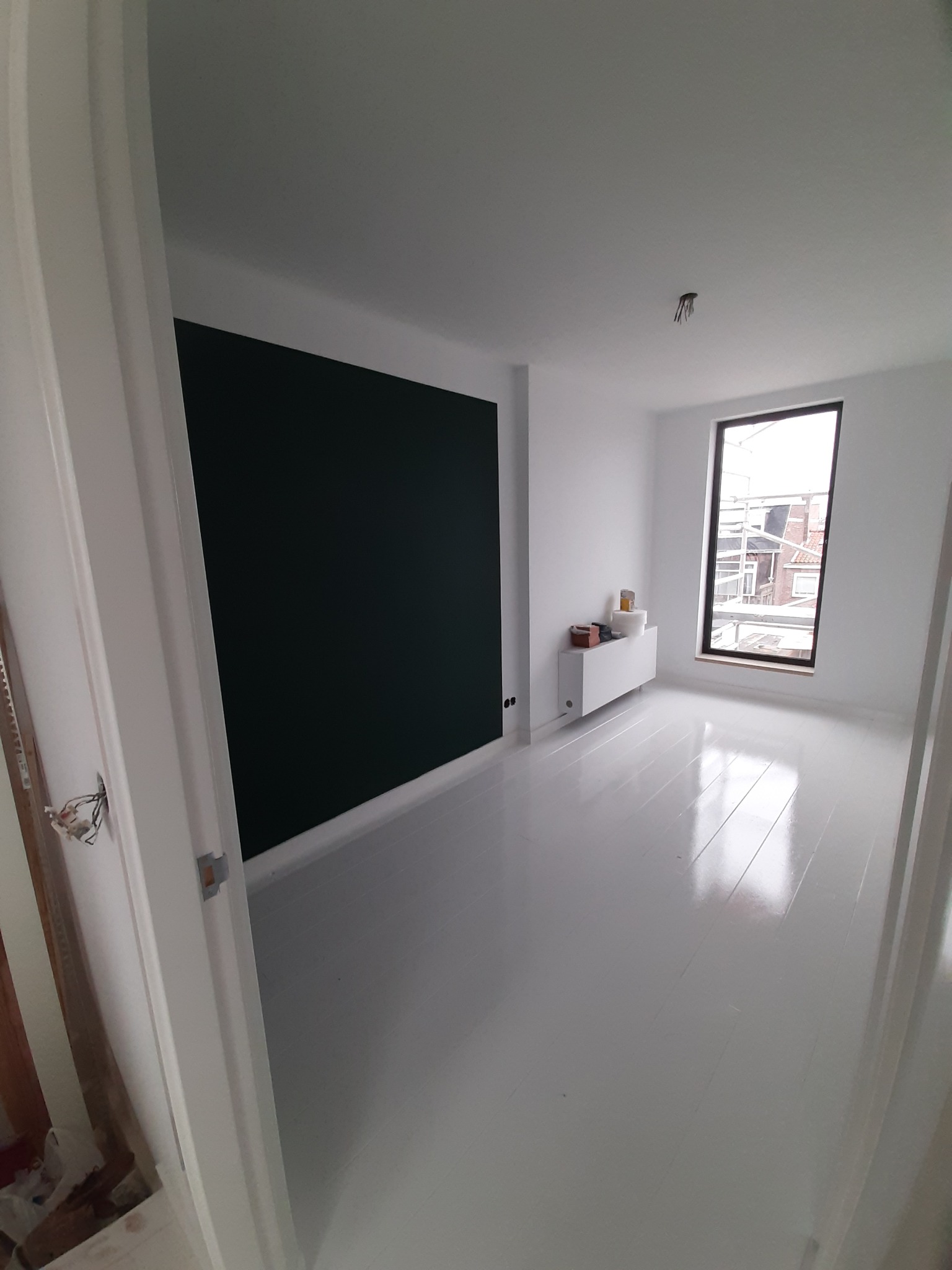 Pomieszczenie po malowaniu: białe ściany, podłoga i stolarka, jedna ściana ciemnozielona. Widoczne puszki z farbą i niedokończone wykończenie przy drzwiach.